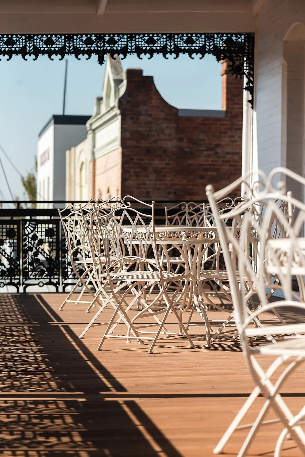 Empty patio with white metal chairs casting shadows on a wooden floor, overlooking neighboring brick and white buildings under clear sky.