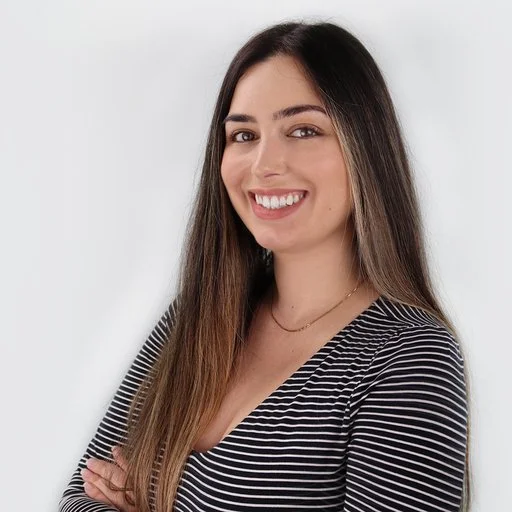 A young woman with long brown hair smiling, wearing a black and white striped shirt and a gold necklace, standing against a white background.
