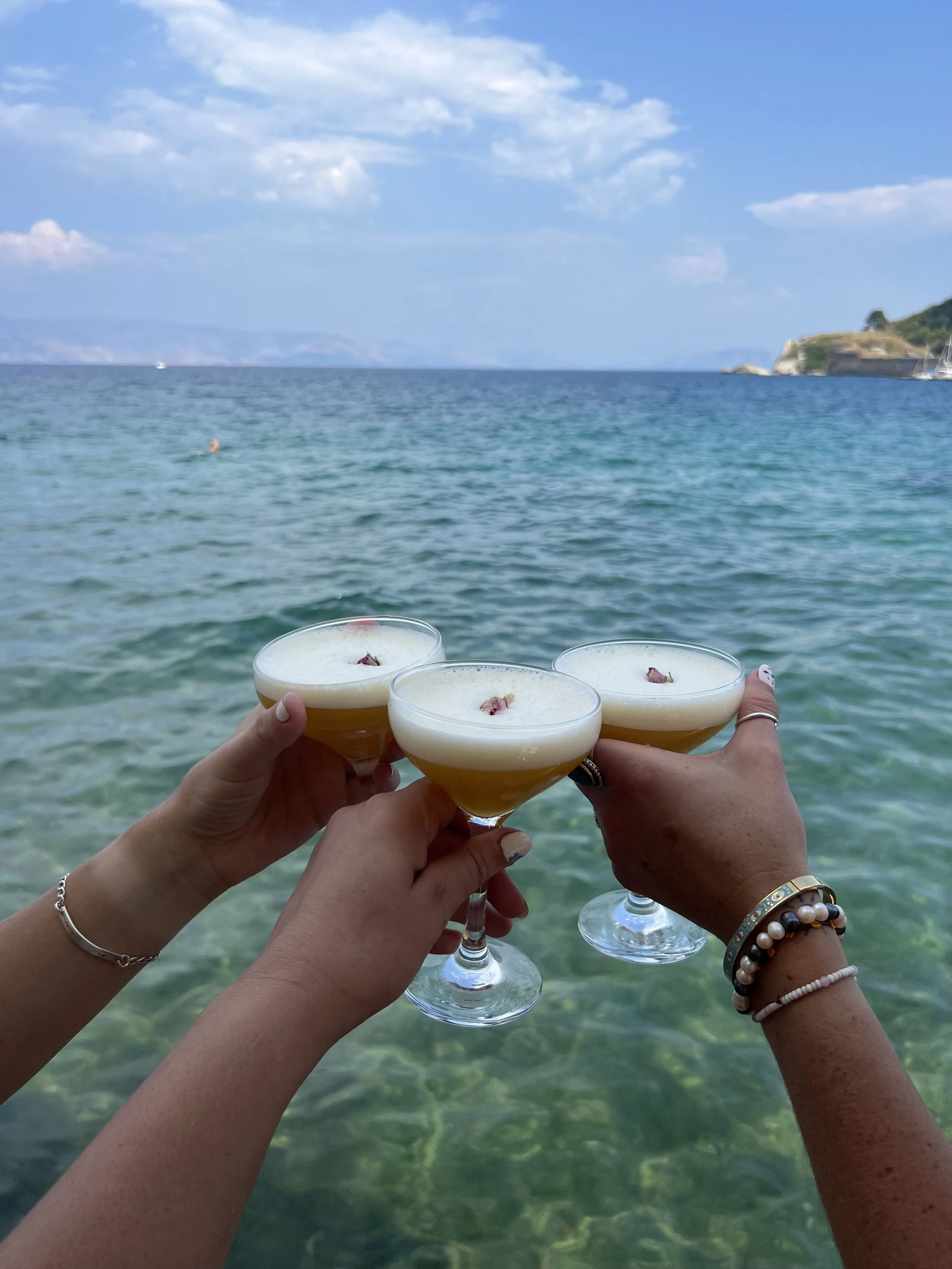 Three people toasting with cocktails over a turquoise ocean with a boat and land in the background.