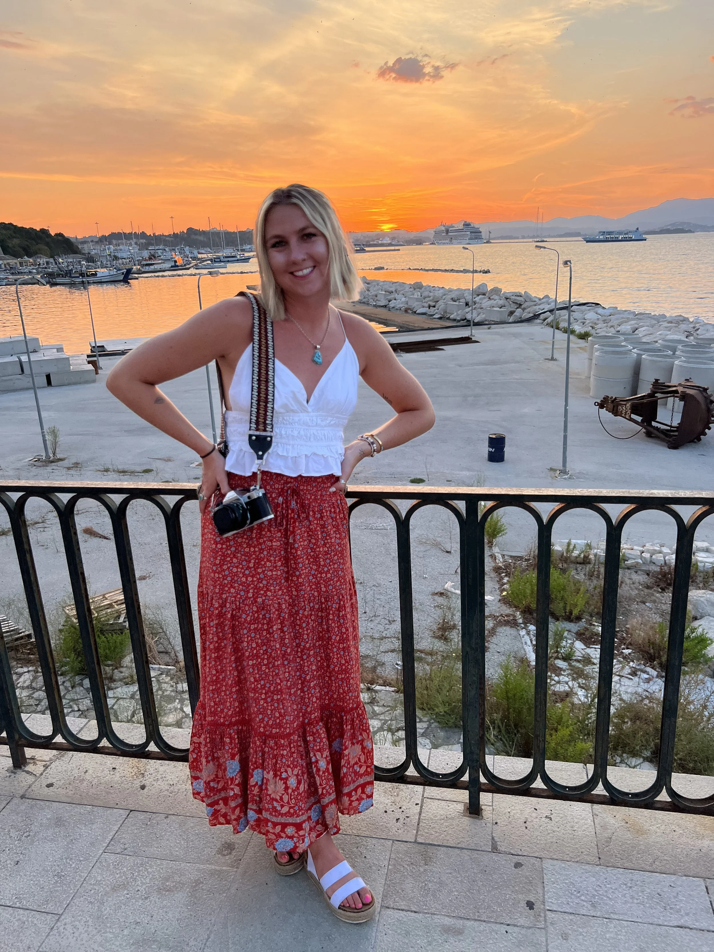 A young woman with blonde hair smiling at the camera, standing in front of a marina at sunset, wearing a white sleeveless top, a long red skirt with floral pattern, white sandals, and carrying a camera with a woven strap.