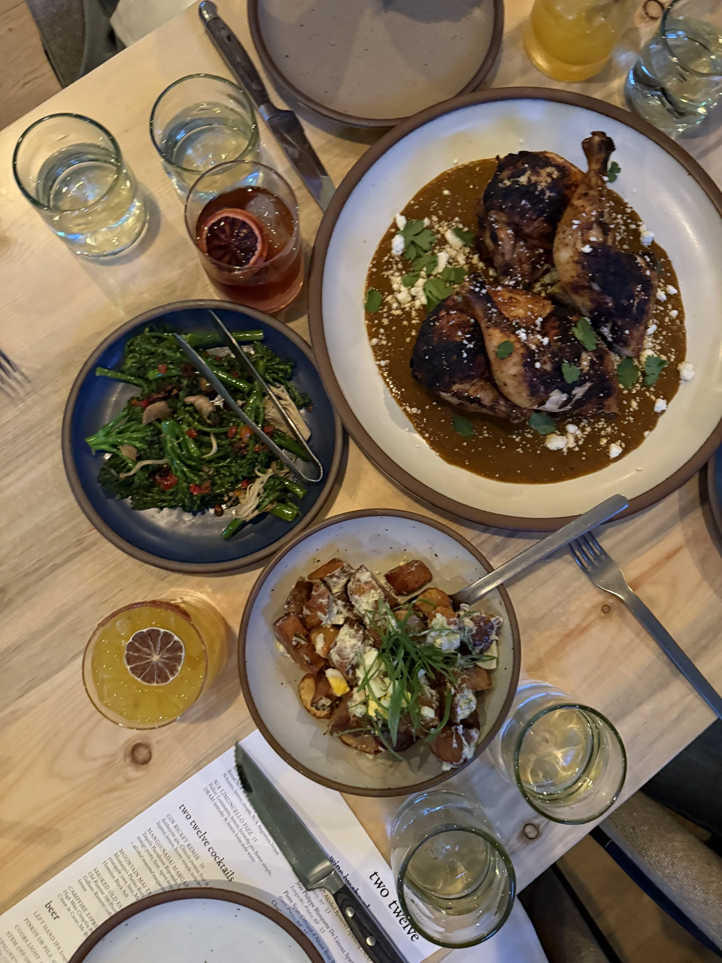 A table with three glasses of water, a glass of iced tea with lemon slice, a large plate of chicken with sauce, a bowl of green vegetables, and a bowl of roasted vegetables with sauce, along with utensils and a menu.
