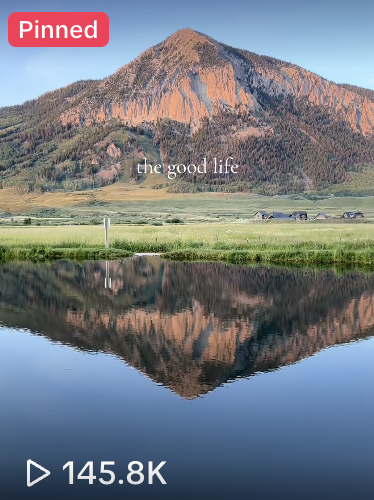 Scenic view of a mountain with a reflection in a calm lake, green fields at the base, and the text 'the good life' overlaid.
