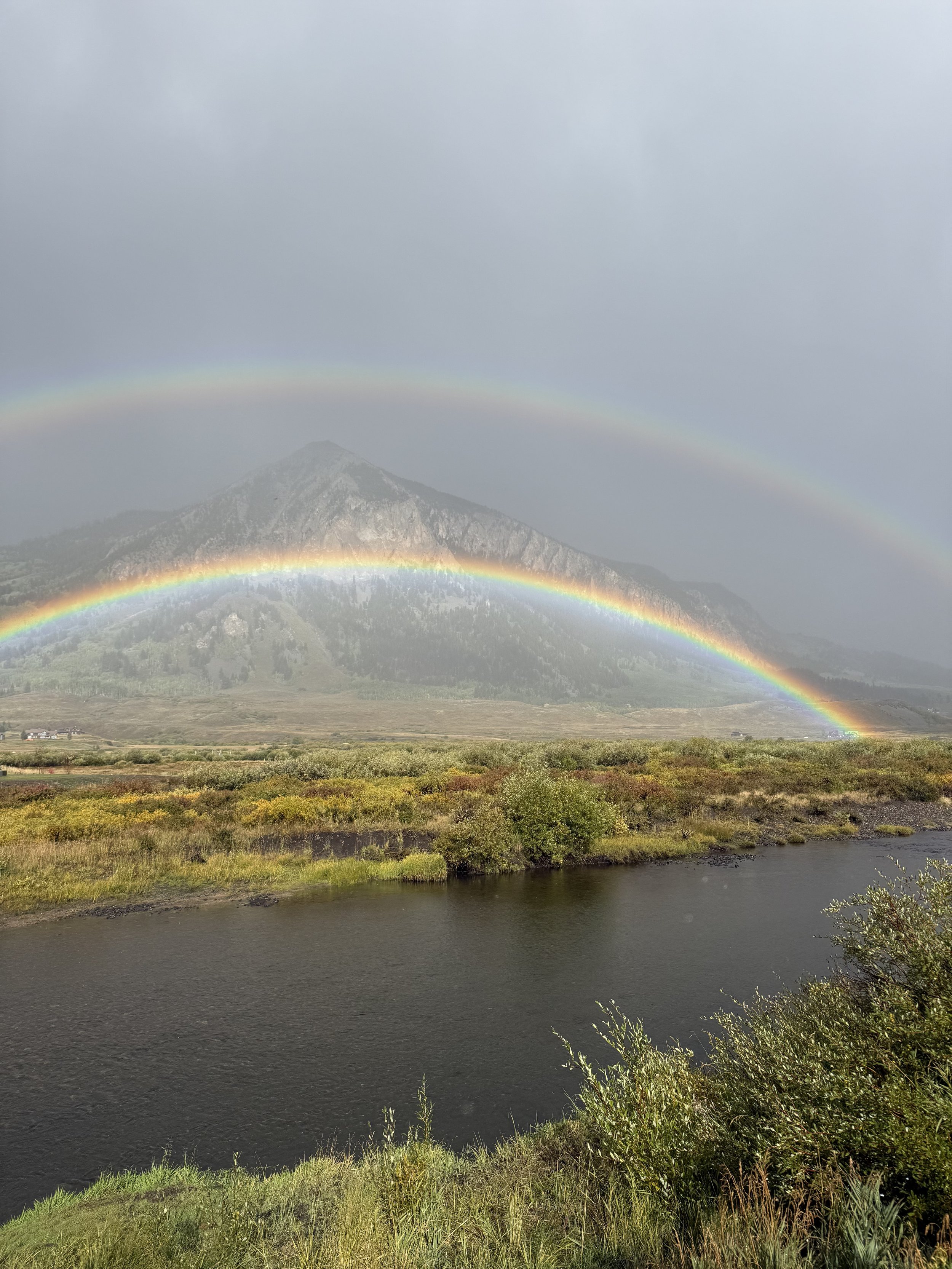 A scenic landscape with a river in the foreground, colorful wetlands, and a mountain in the background under a cloudy sky. Two rainbow arcs are visible in the sky.