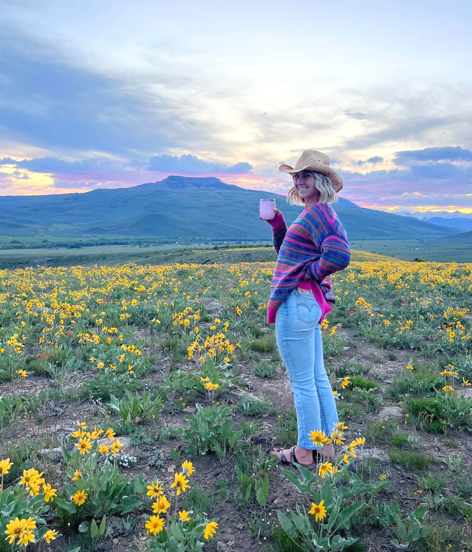 A woman in a colorful striped sweater, light blue jeans, and a straw hat stands in a field of yellow flowers at sunset, holding a cup and smiling, with mountains in the background.