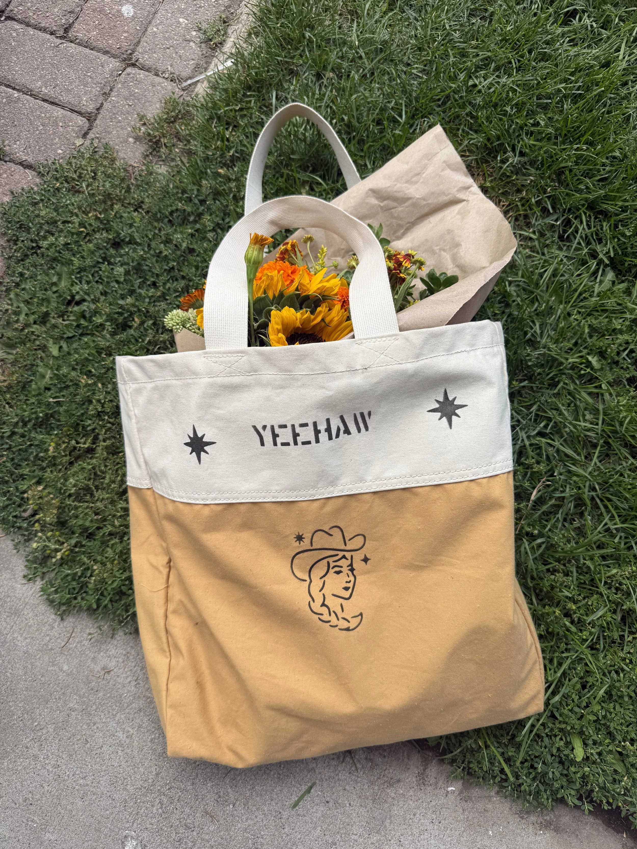 A tote bag with a yellow and white design, featuring the word 'YEEHAA' and a line drawing of a woman with a hat, lying on grass with a bouquet of orange and yellow flowers inside.