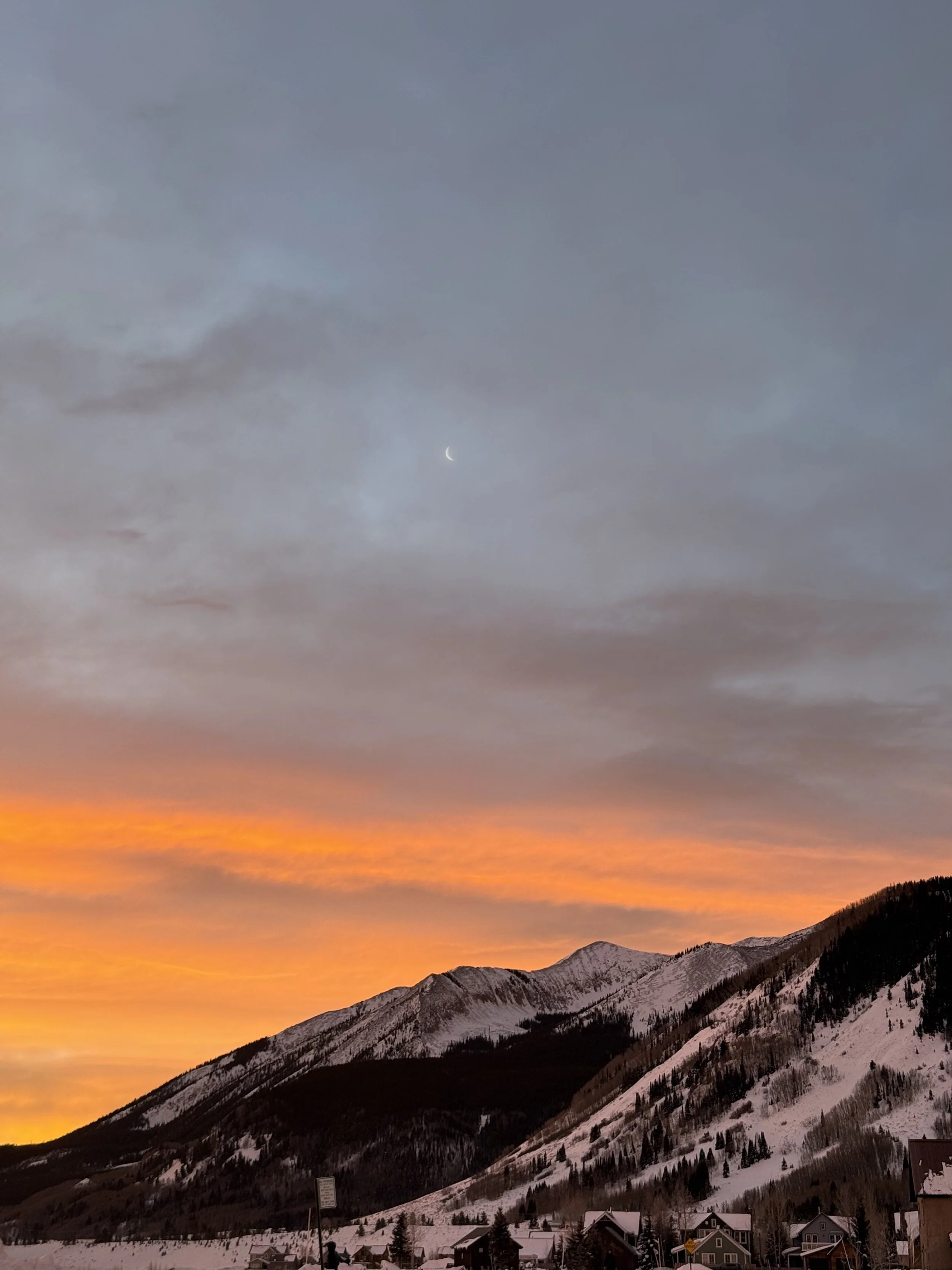 Snow-covered mountains at sunset with orange and yellow sky, clouds, a crescent moon, and houses at the base.