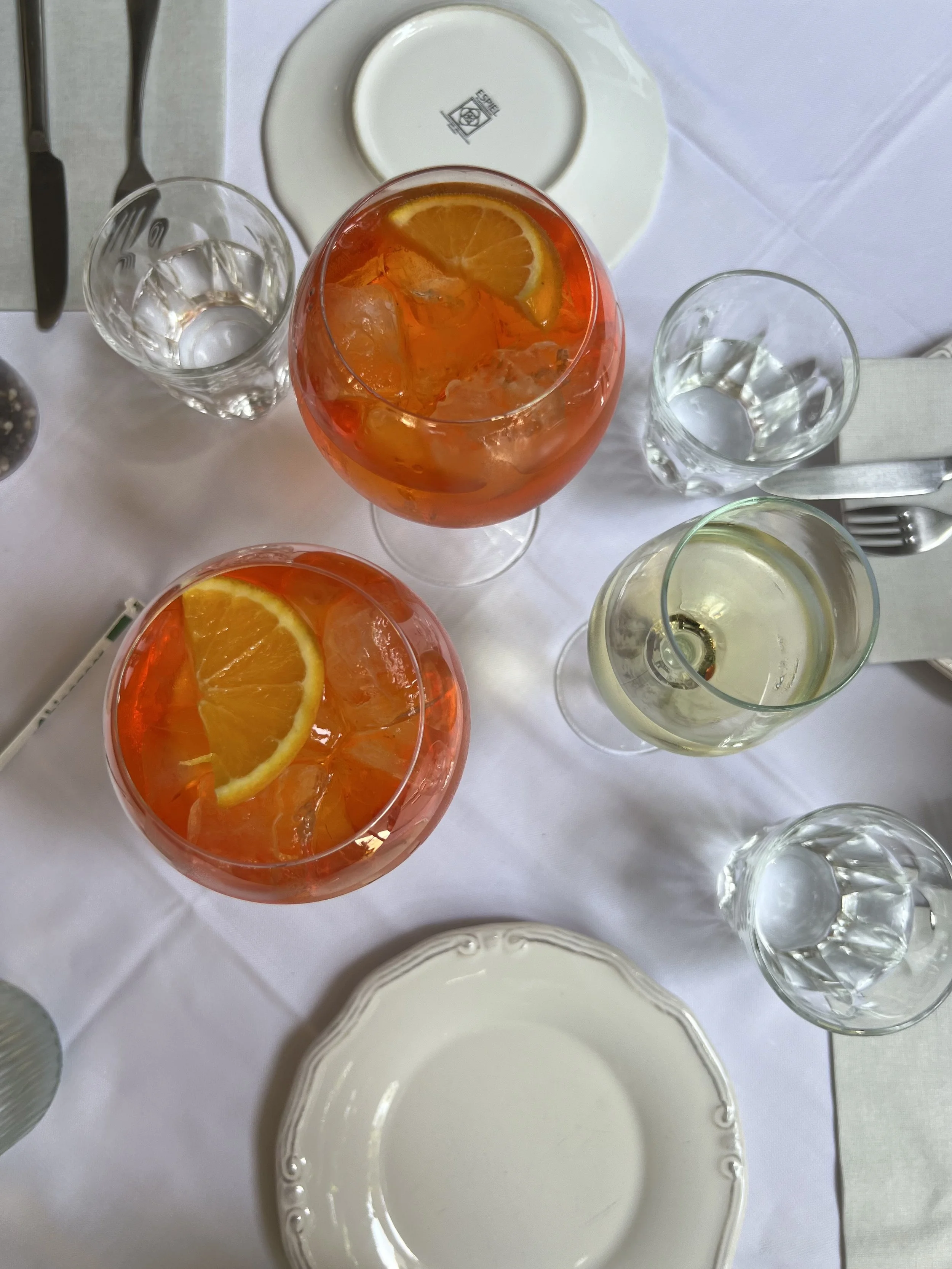 A top-down view of a table setting with two glasses of orange-colored cocktails with lemon slices, a glass of white wine, several empty glasses, a white plate with a logo on the back, and silverware on a white tablecloth.