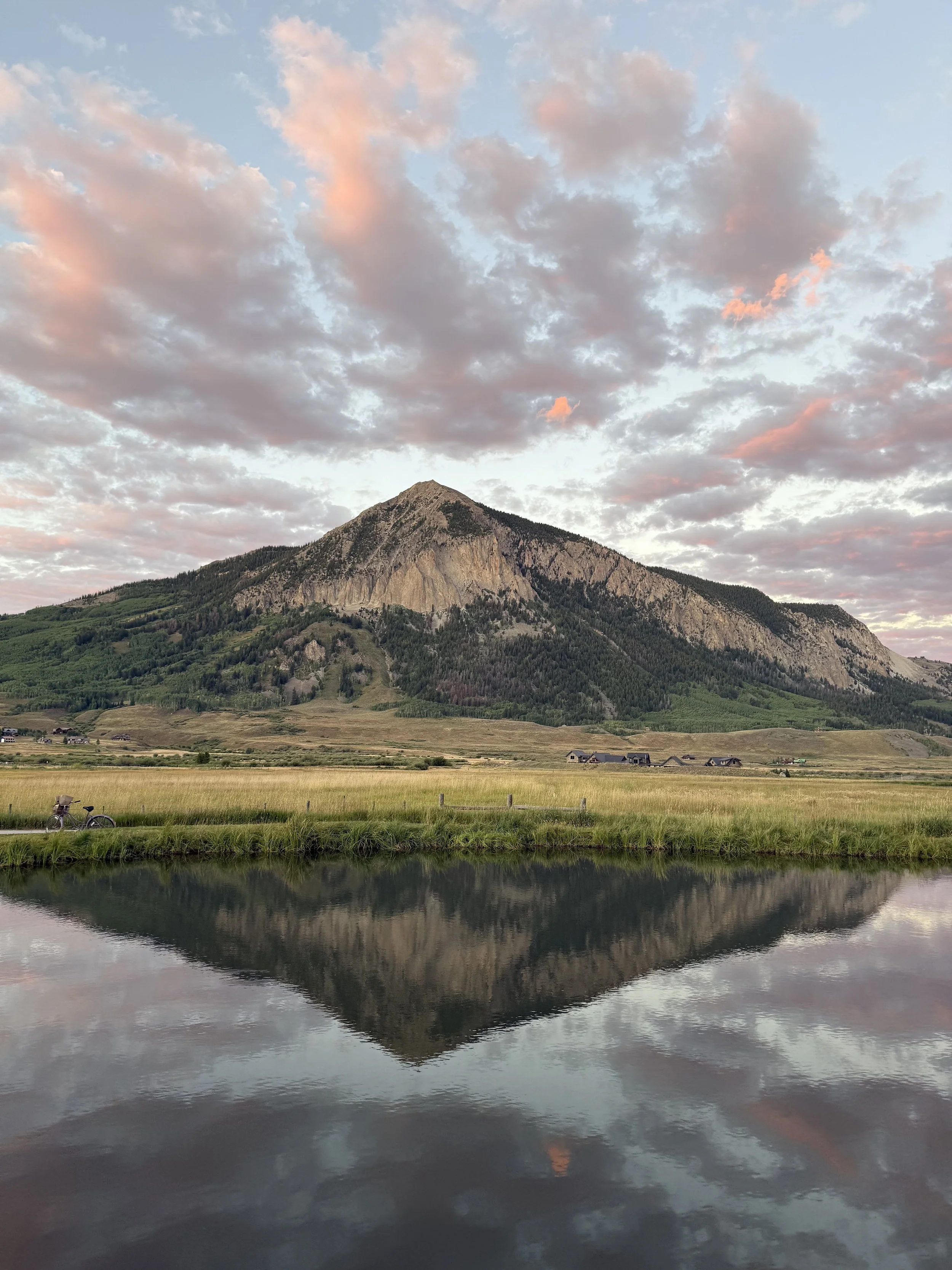 Scenic view of a mountain with a reflection in the calm water in the foreground, cloudy sky with pink hues at sunset, green fields, and a bicycle on the grass.