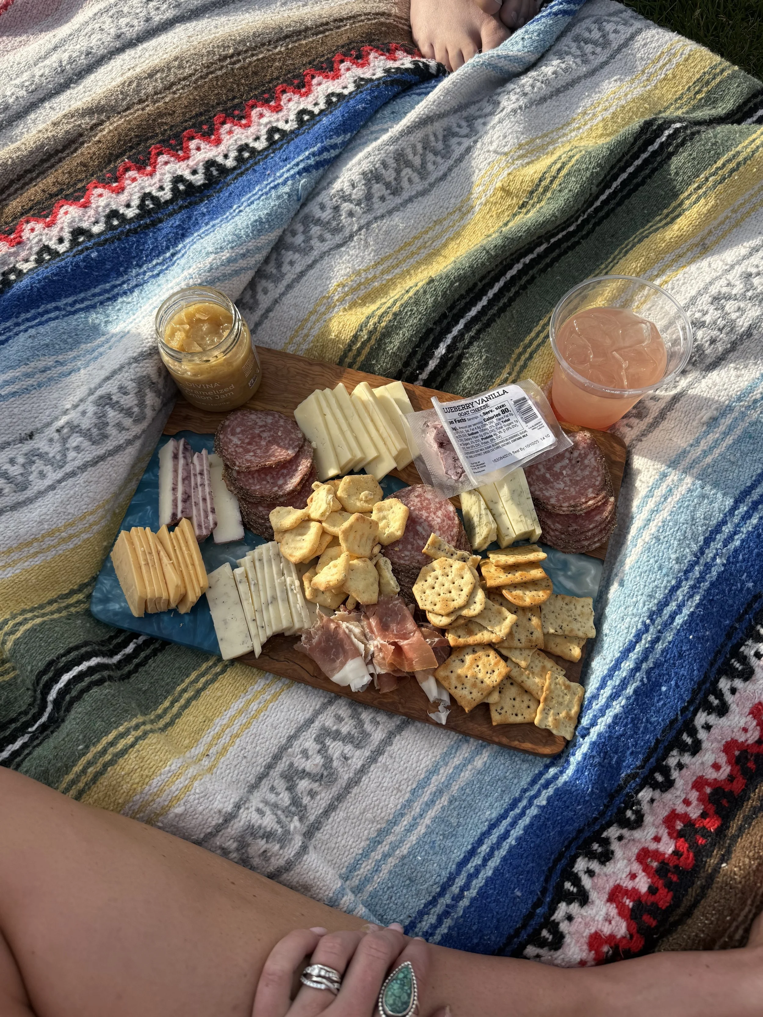Picnic with a cheese and charcuterie board on a colorful striped blanket, with a drink and a jar of honey.