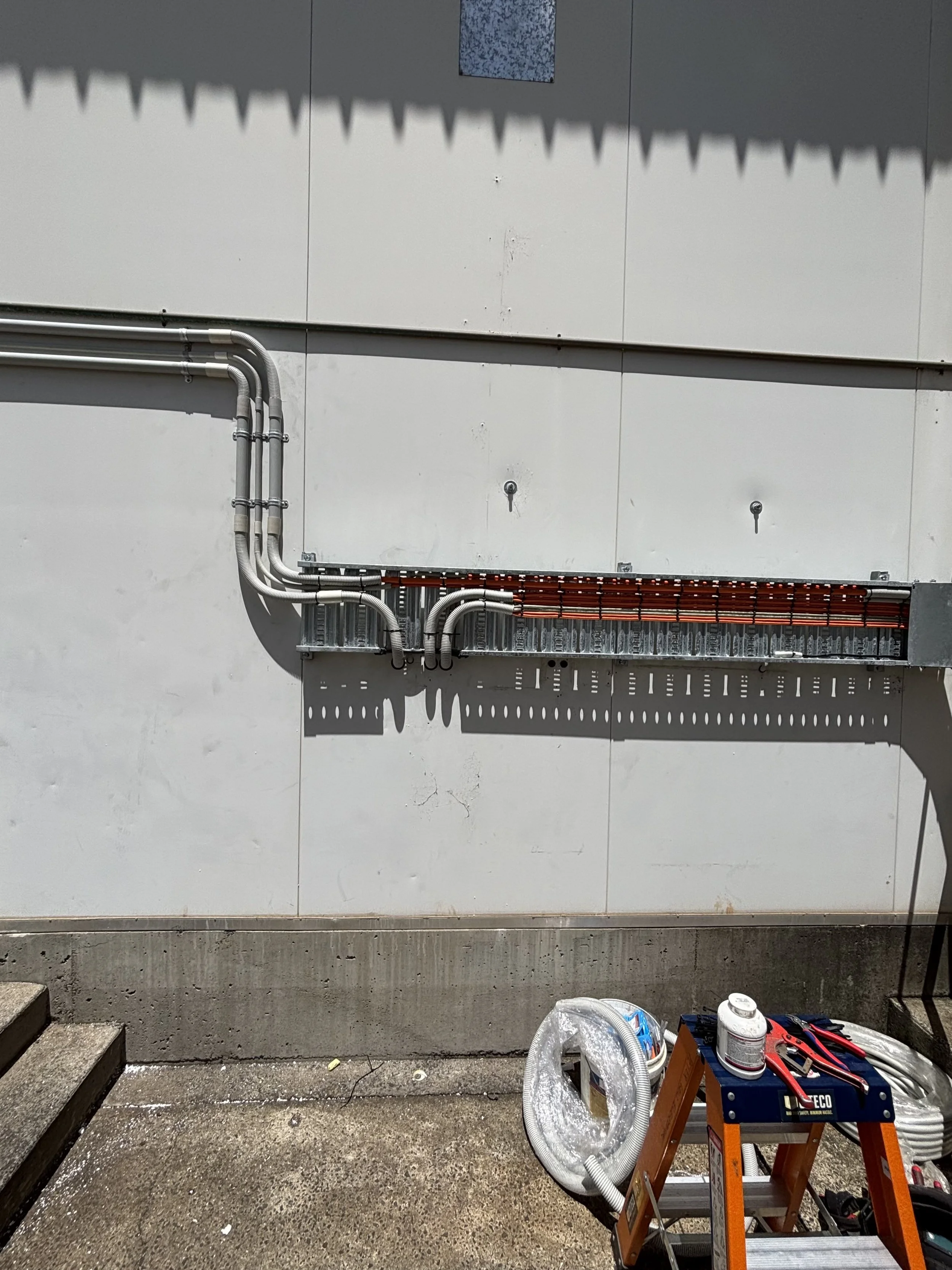 Electrical wiring and conduits installed on an exterior wall, with construction tools and materials on the ground below.