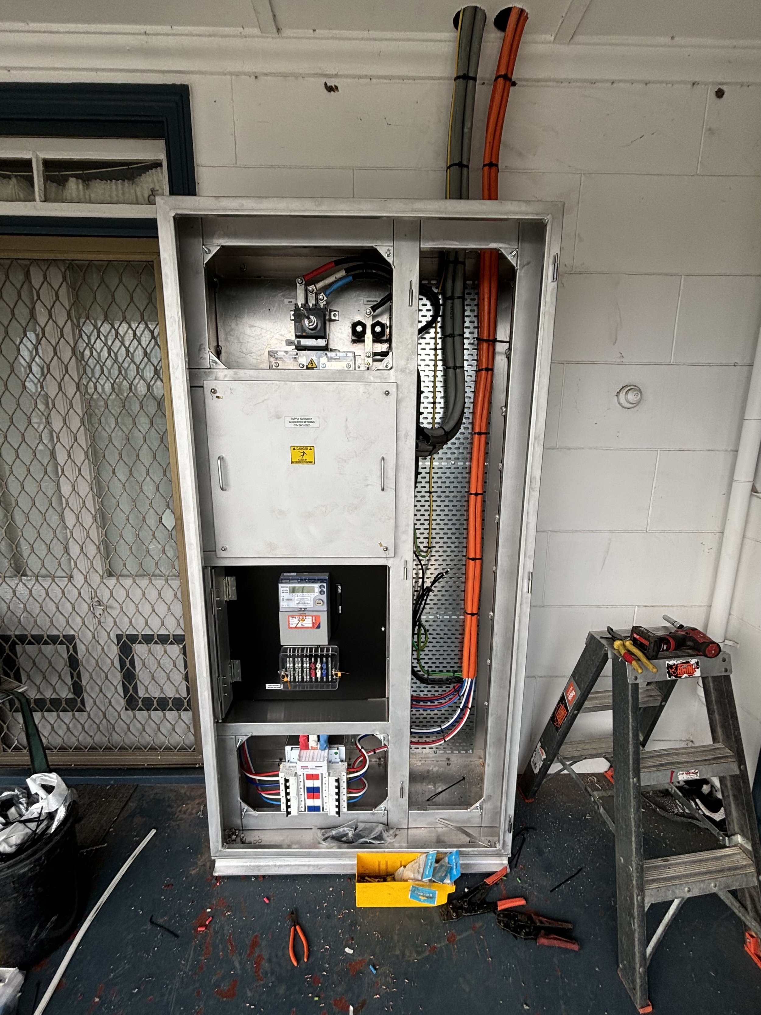 An electrical meter and components inside a metal enclosure with tools and wires nearby, in a workshop or utility room.