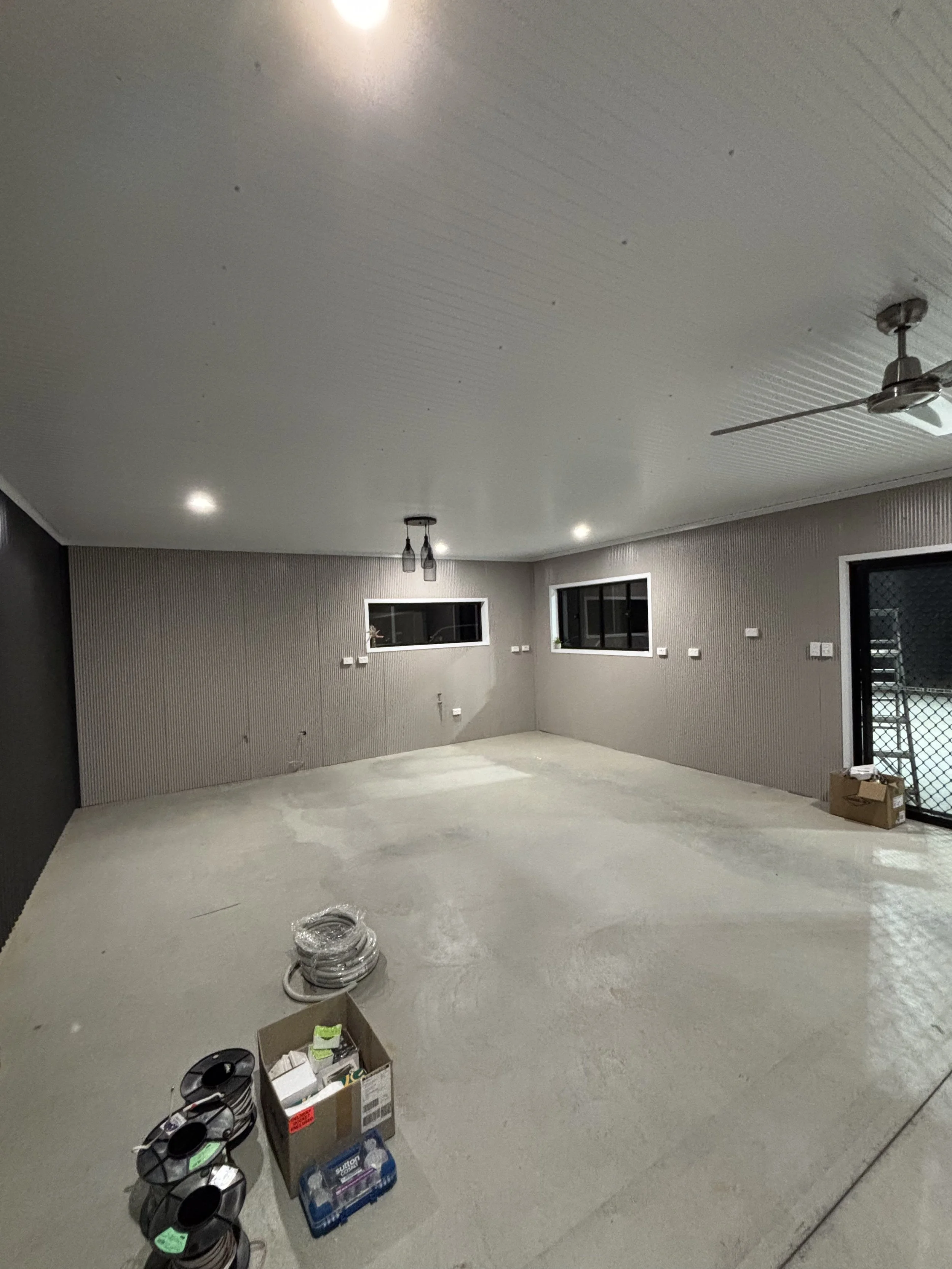 An empty, unfinished room with a concrete floor, gray textured walls, and a white ceiling. There are two small horizontal windows, a ceiling fan, and pendant lights. A box and spools of wire are on the floor near the doorway.