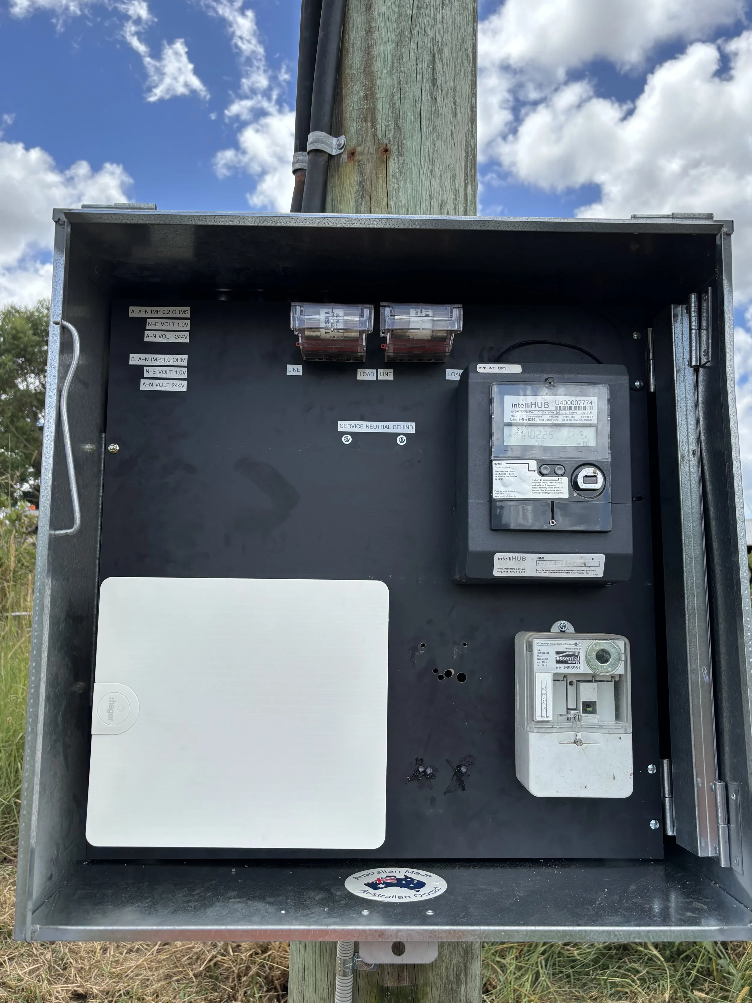 An open electrical or utility box mounted on a pole, containing a digital meter, switches, and wiring, with a partly cloudy sky in the background.