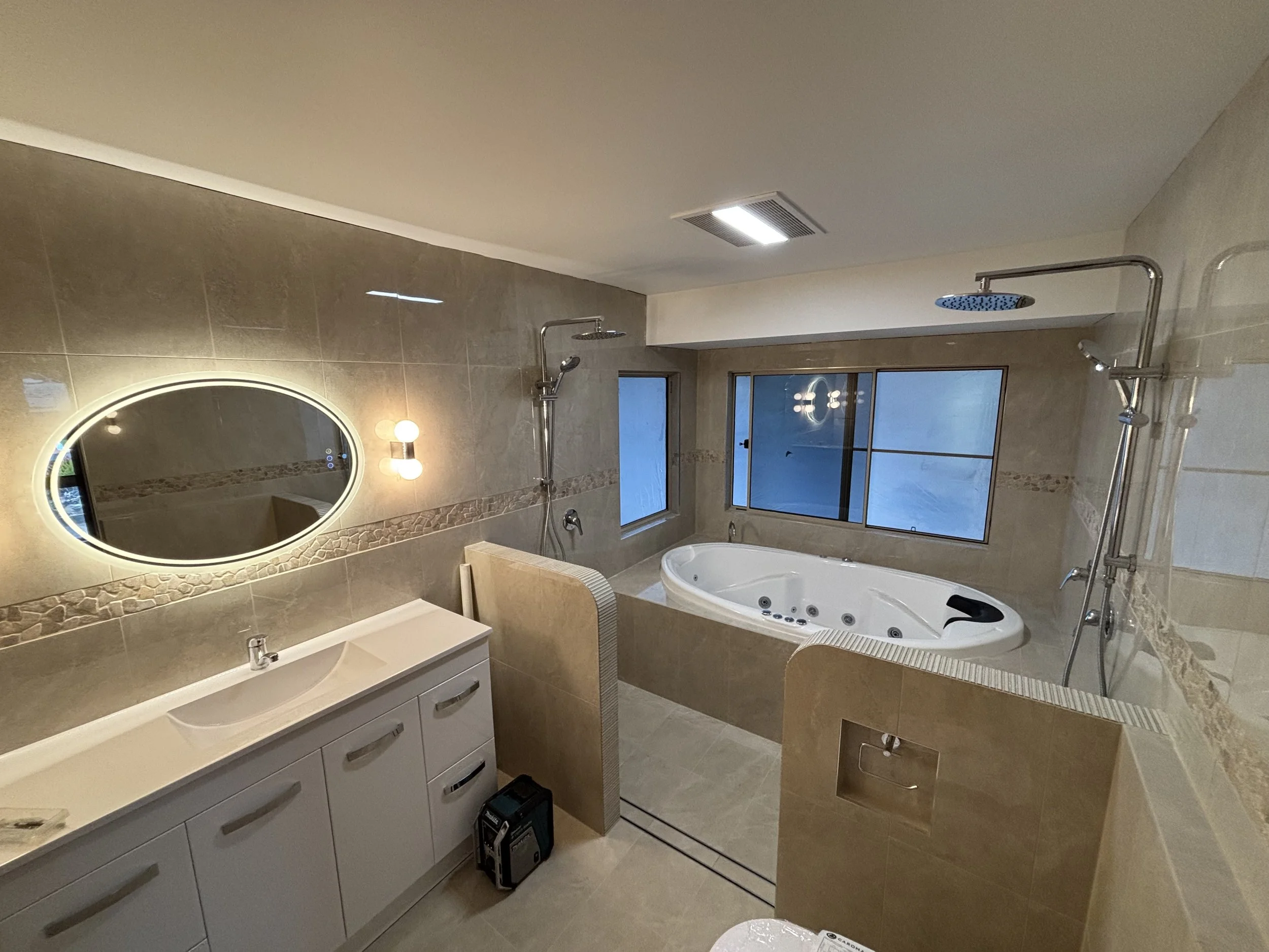 Modern bathroom with a large whirlpool bathtub, dual rainfall showers, a white vanity with a curved sink, an oval backlit mirror, a wall-mounted light fixture, and a frosted window.