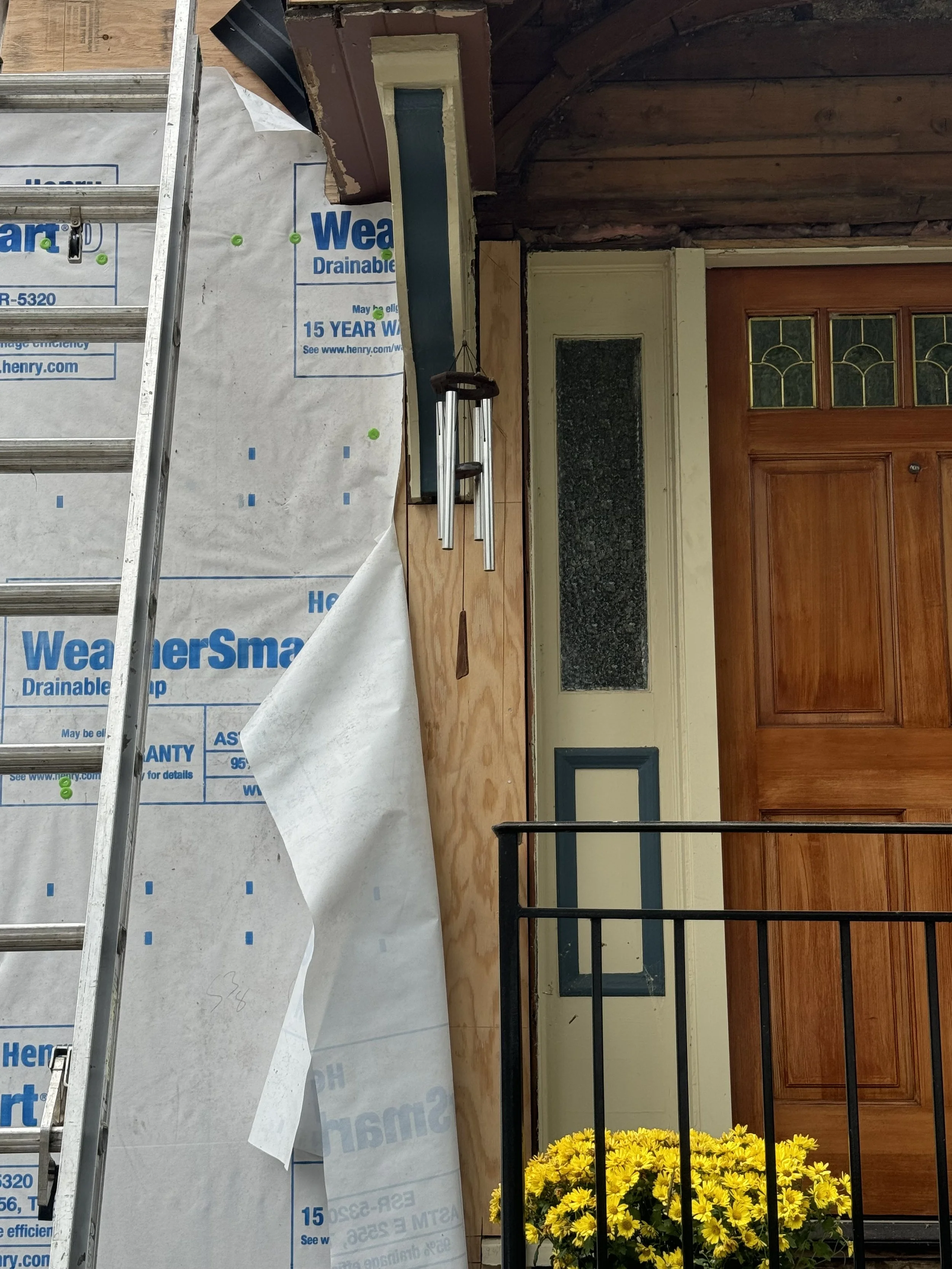 Construction or renovation work on the front door of a house, with a ladder and weather barrier covering part of the exterior wall, which has a wooden door with stained glass and a yellow flower pot in front.
