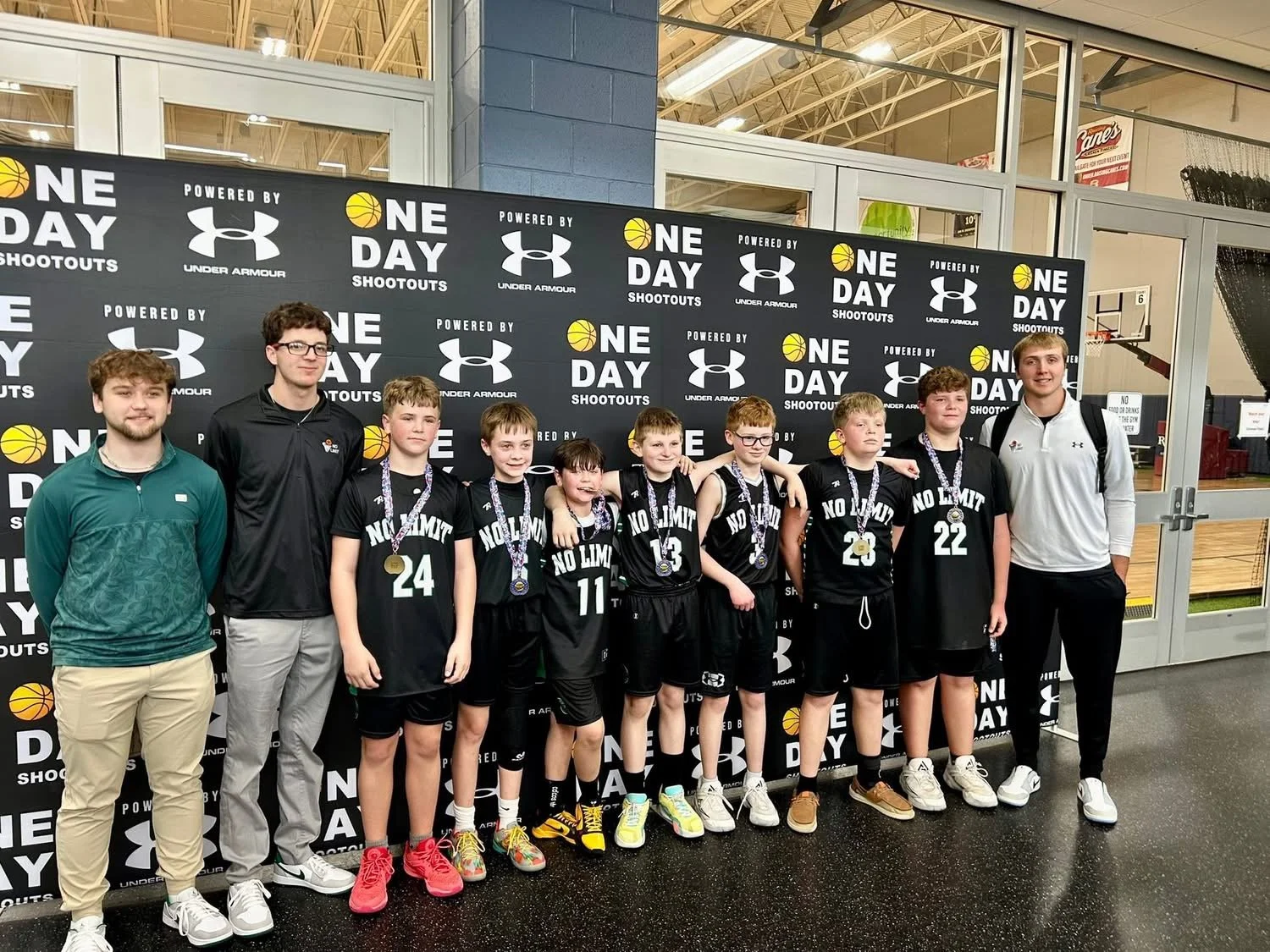 Group of young boys and two coaches at a basketball event, with medals around their necks, standing in front of a black backdrop with logos and the text 'One Day Shootouts' and 'Powered by Under Armour'.