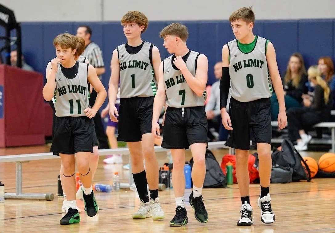 Four young boys wearing basketball jerseys with 'NO LIMIT' printed on them, standing on a basketball court, with spectators in the background.