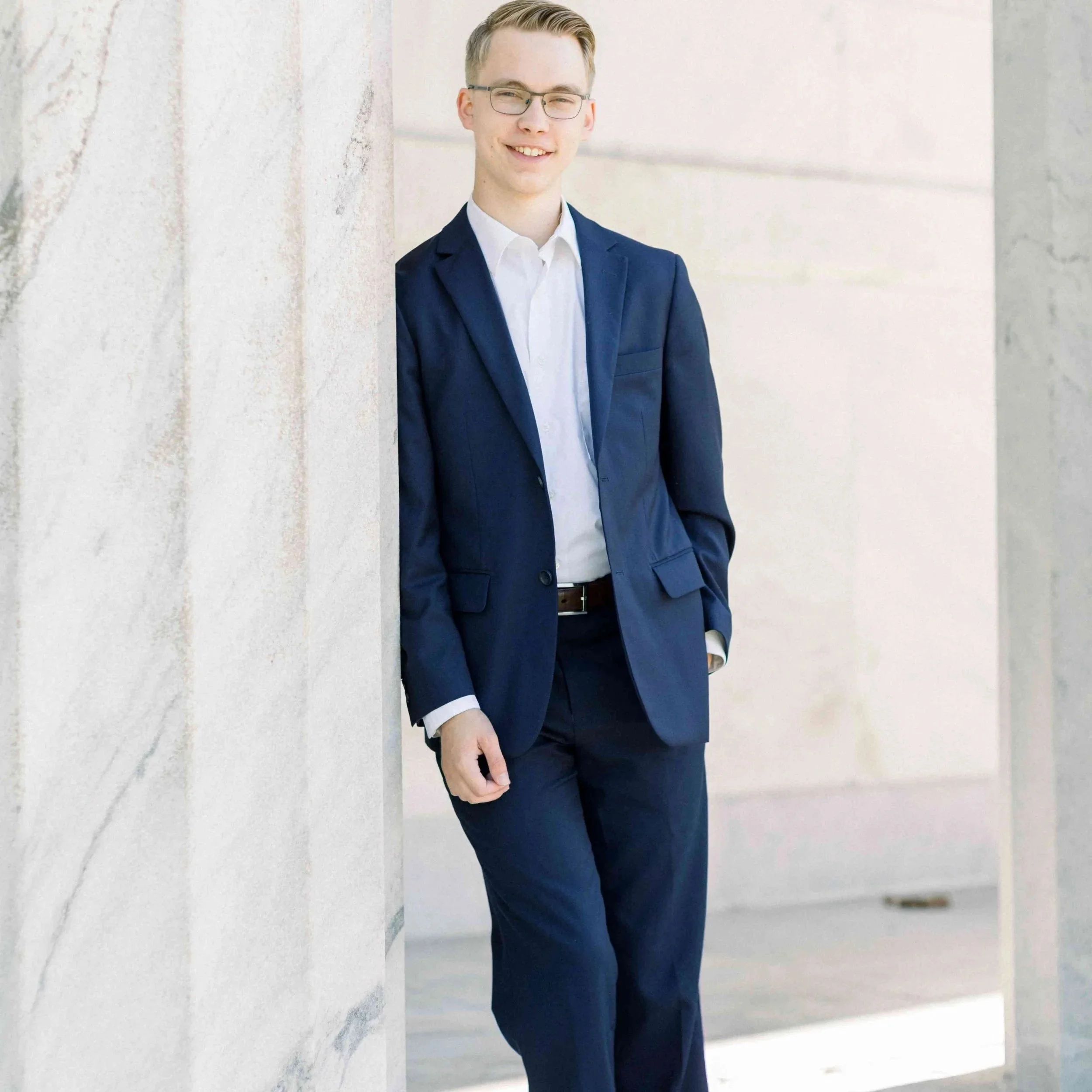 A young man in a navy blue suit and white shirt leans against a light-colored wall, smiling.