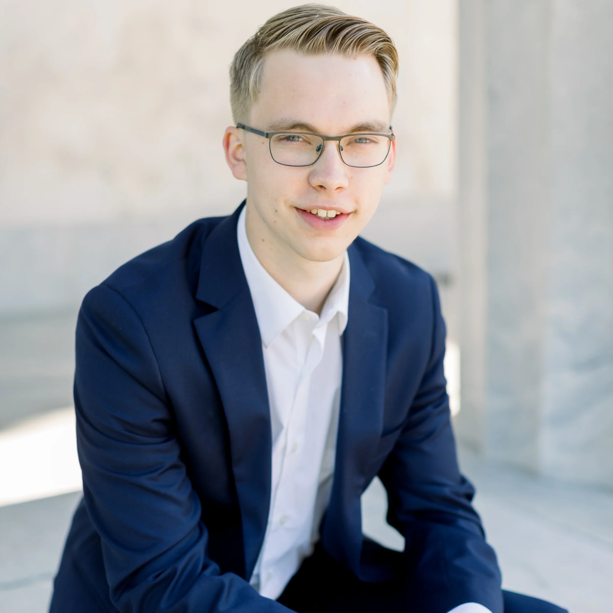 Portrait of a young man in a navy blazer and white shirt, wearing glasses, sitting outdoors with a blurred stone background.