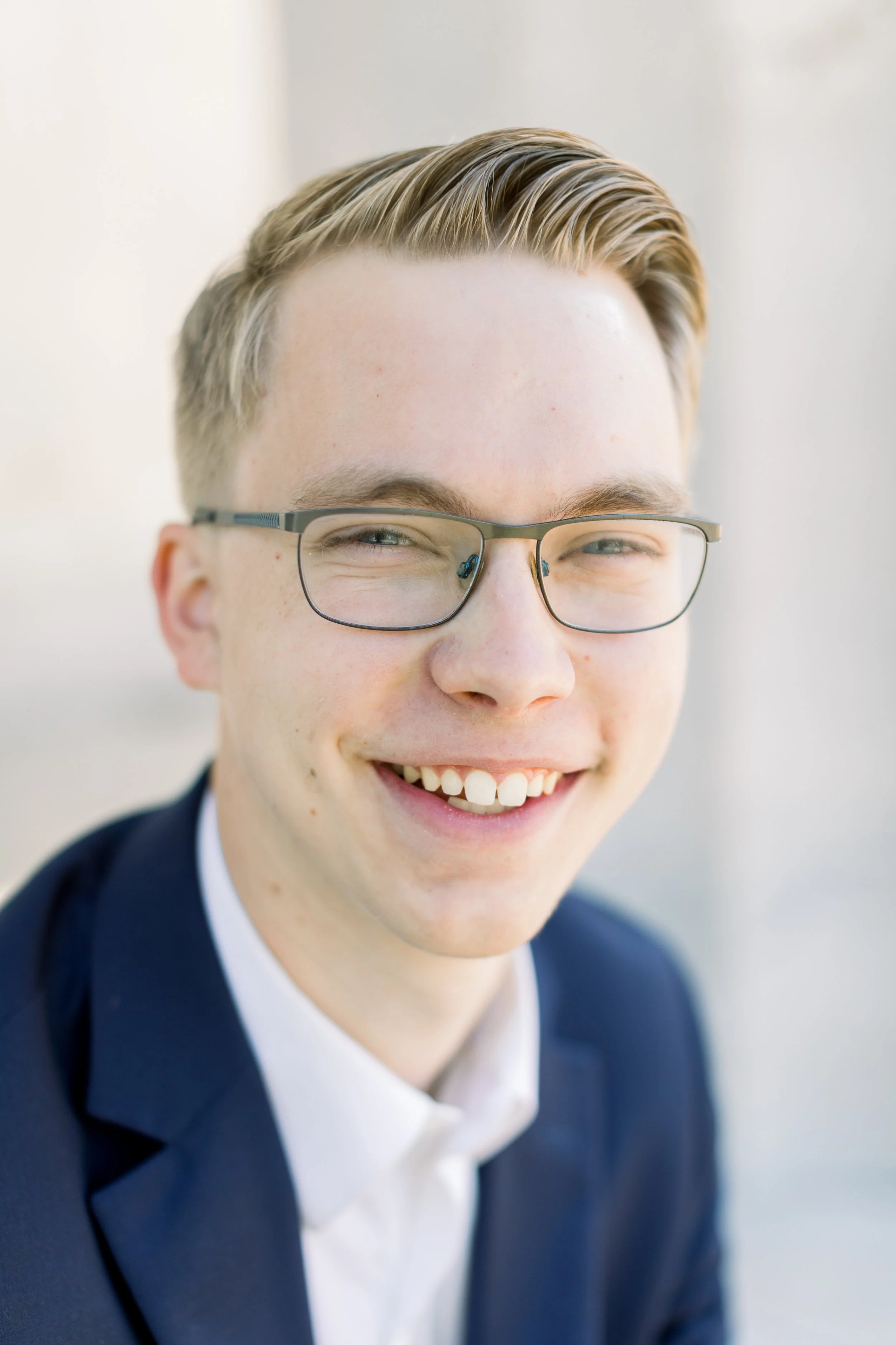 Close-up of a young man smiling, wearing glasses and a navy blazer, with blonde hair and a white shirt, outdoors with a blurred background.