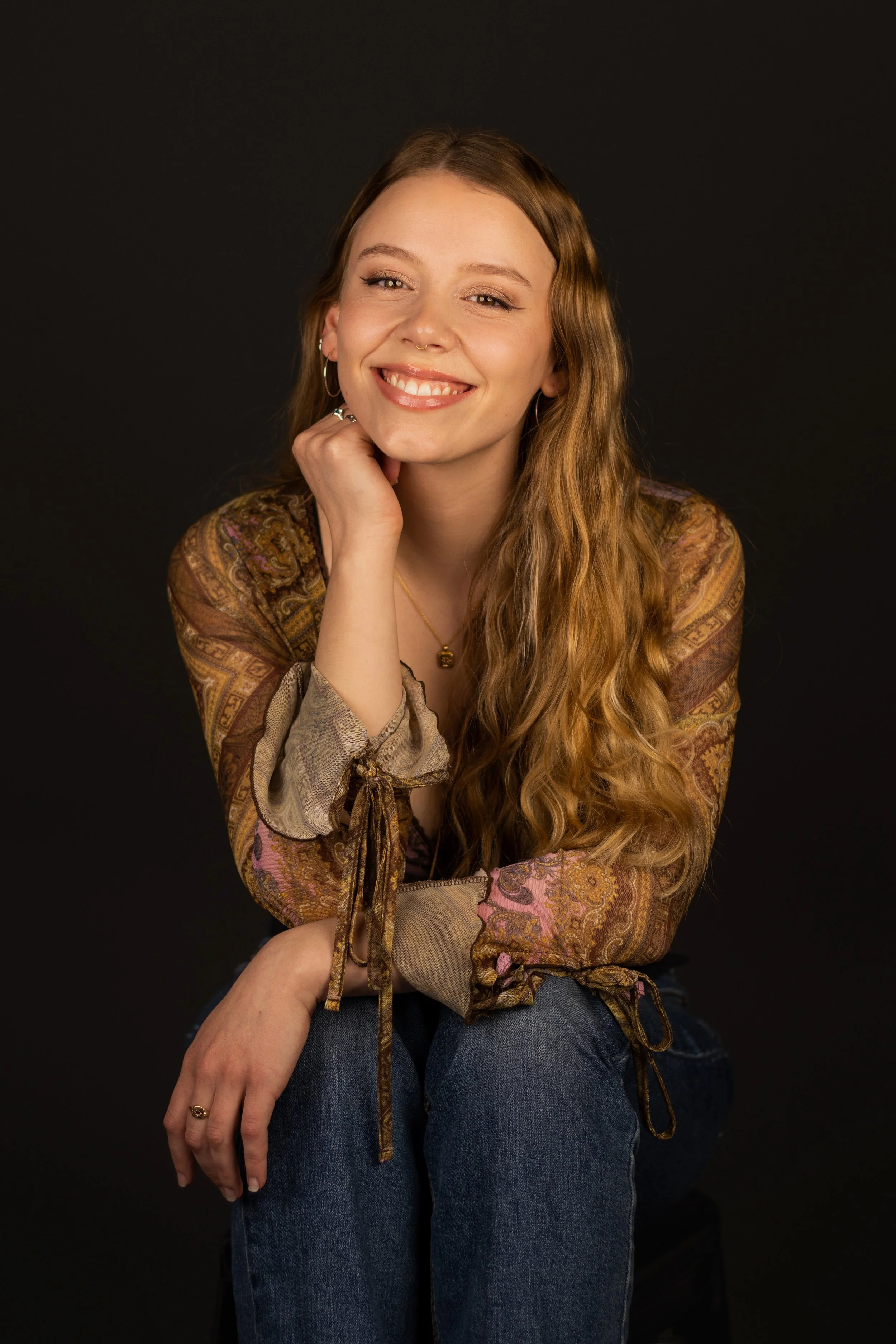 A young woman with long wavy hair, smiling and resting her chin on her hand, sitting against a black background. She is wearing a patterned blouse, blue jeans, and jewelry including earrings, a ring, and a necklace.