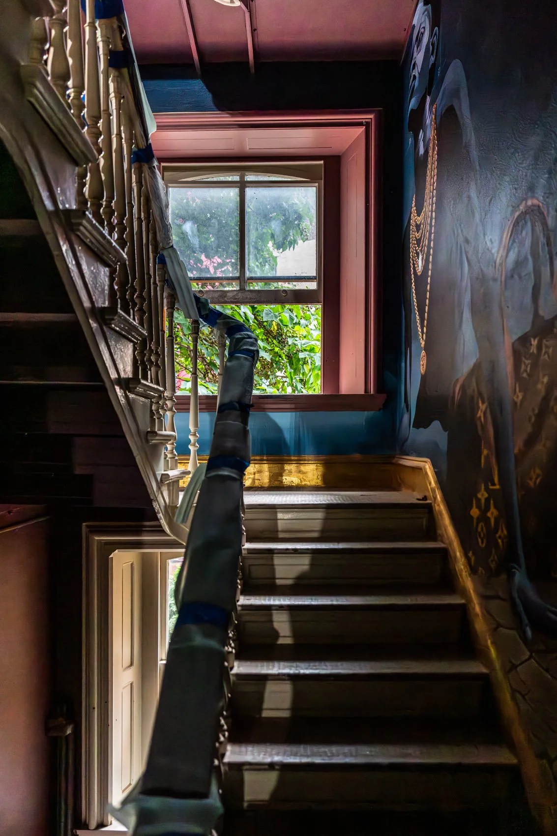Upside-down view of a staircase with a wooden railing, leading to a window with greenery outside, inside a house with ornate wall art.