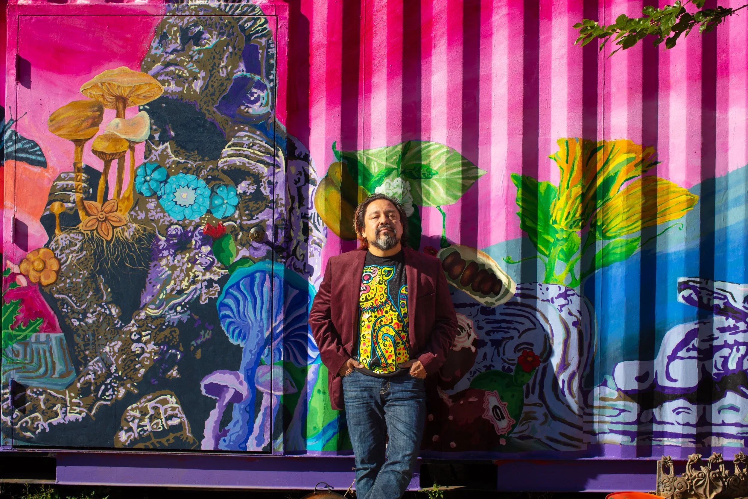 A man with dark hair and a beard, wearing a maroon blazer and a colorful, abstract T-shirt, stands with hands in pockets in front of a vibrant mural depicting large leaves, mushrooms, and abstract faces on a pink and purple background.