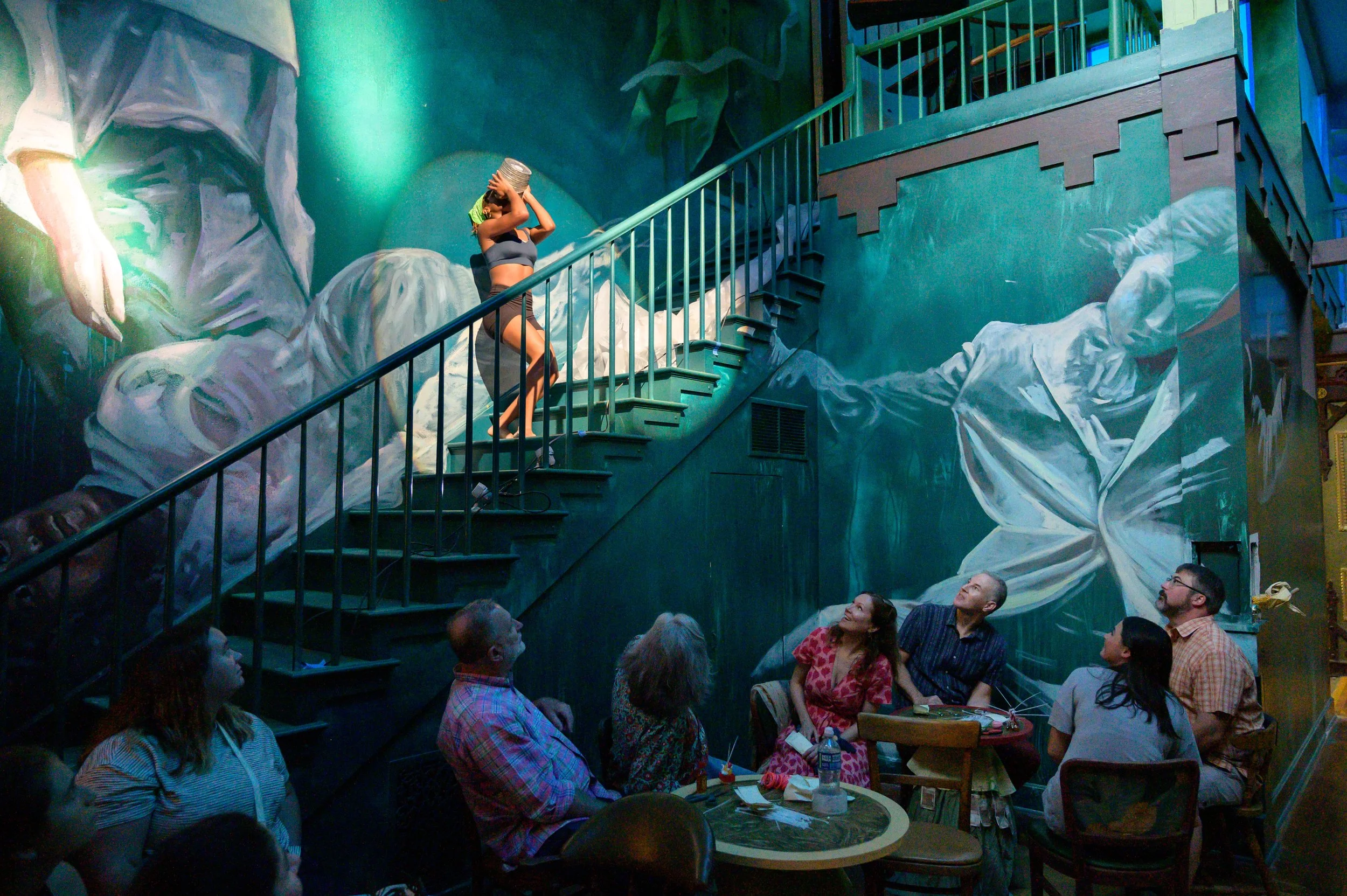 A woman on stage wearing a gray crop top and shorts, with a headpiece, performing for an audience in front of a large, colorful mural with abstract faces and figures.