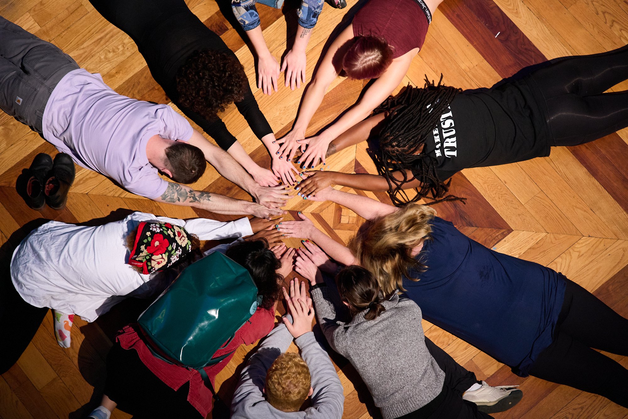 A group of diverse people lying on a wooden floor in a circle, stacking their hands together in the center.