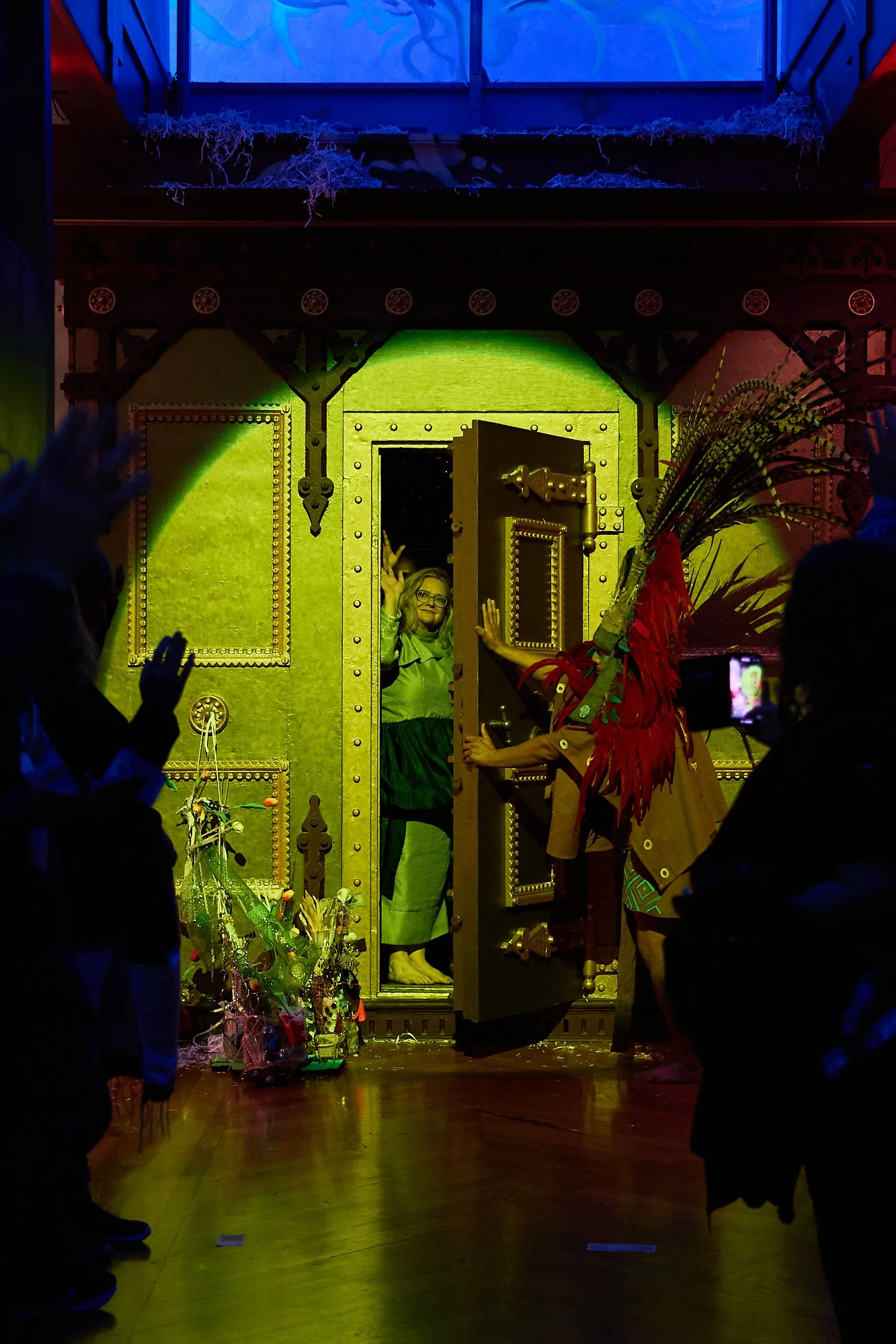 A woman in glasses standing in a large doorway, waving. The doorway is decorated with studs and gold accents, with someone in a colorful costume with feathers and a headdress standing nearby, partially turned away from camera. People are gathered around, some taking photos, in a dimly lit room with colorful lighting.