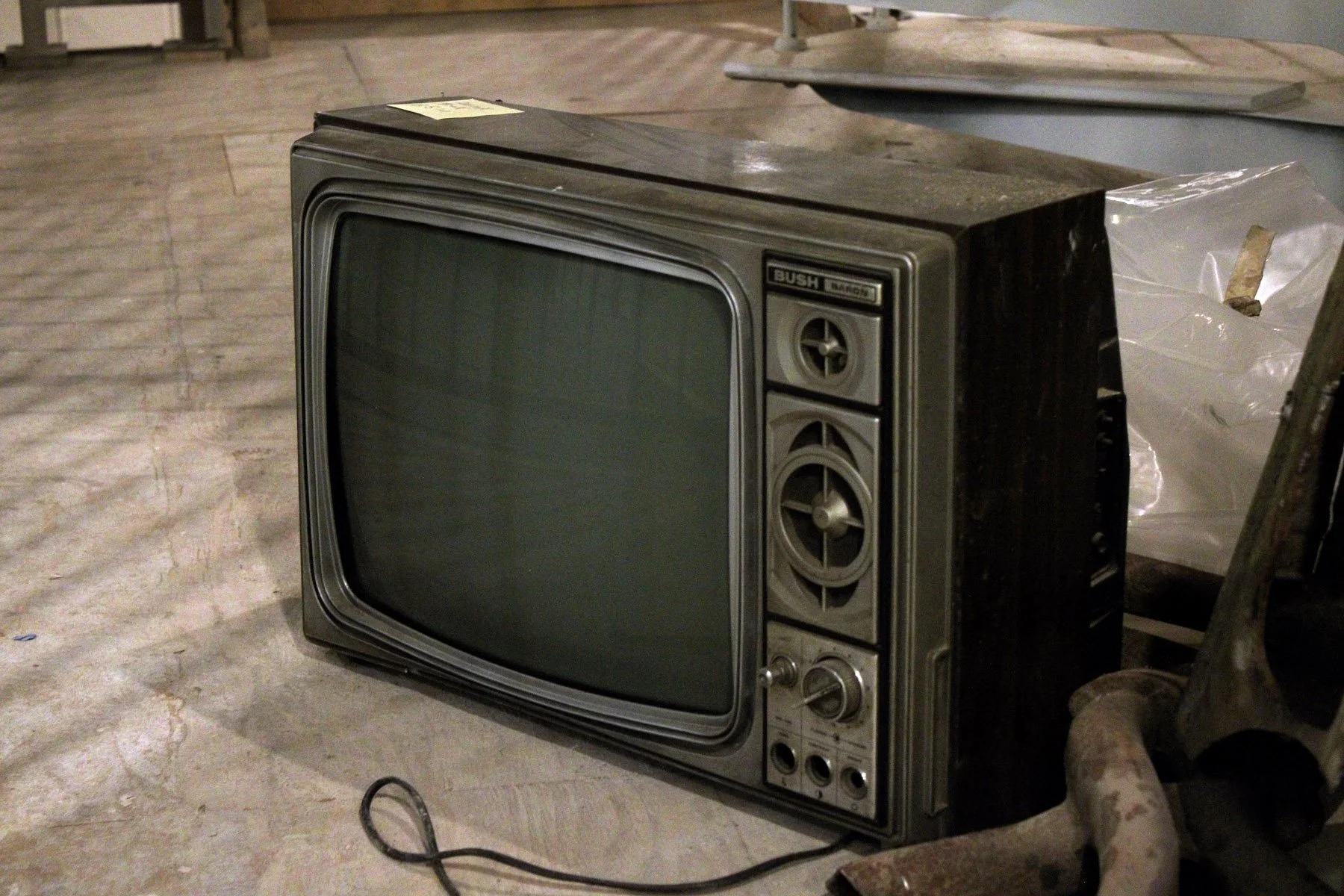 Old, black and silver vintage television from the 1960s or 1970s in a room with a debris-covered floor and a plastic bag nearby.