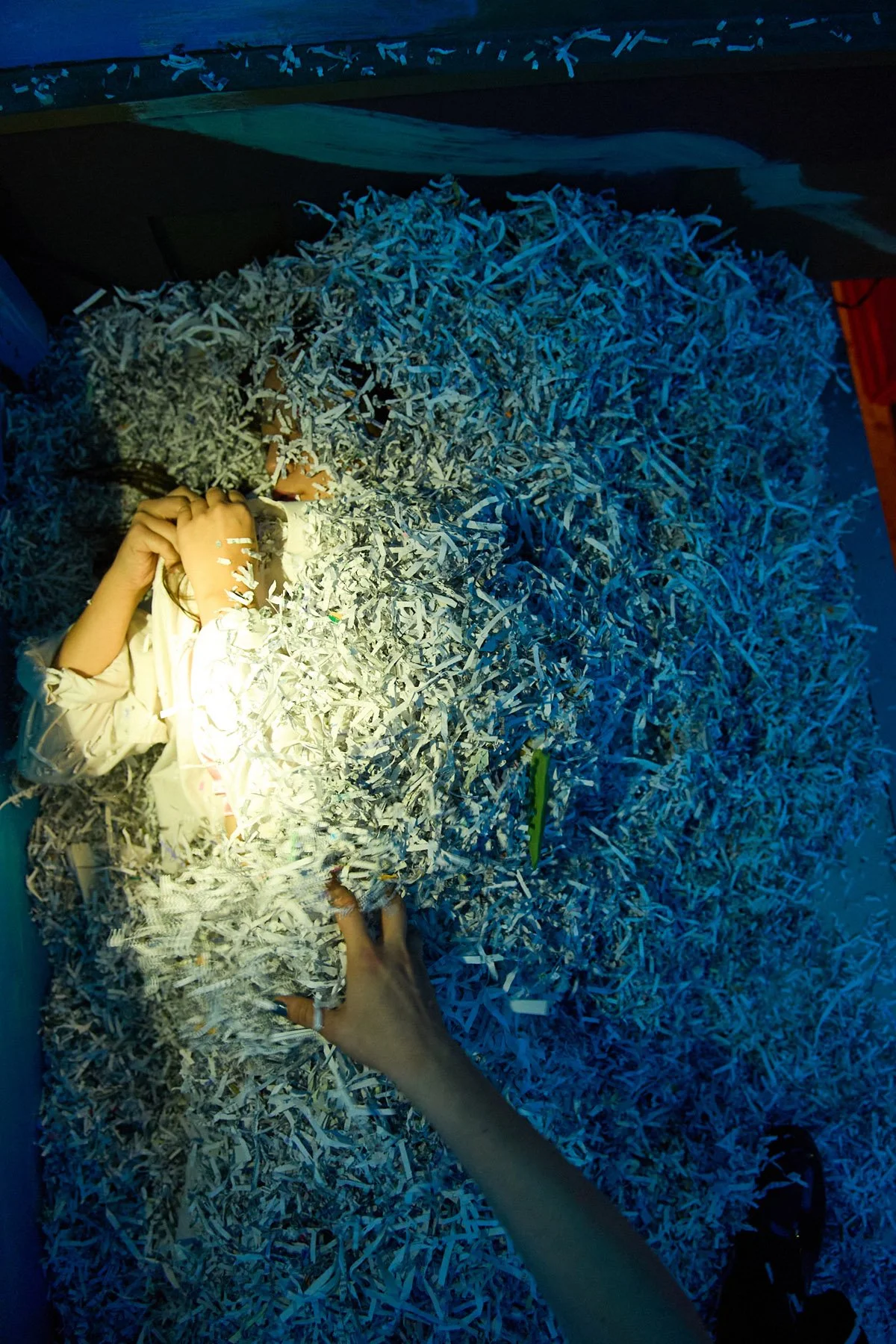 Person lying in a bed of shredded paper with another person reaching in to touch the paper.