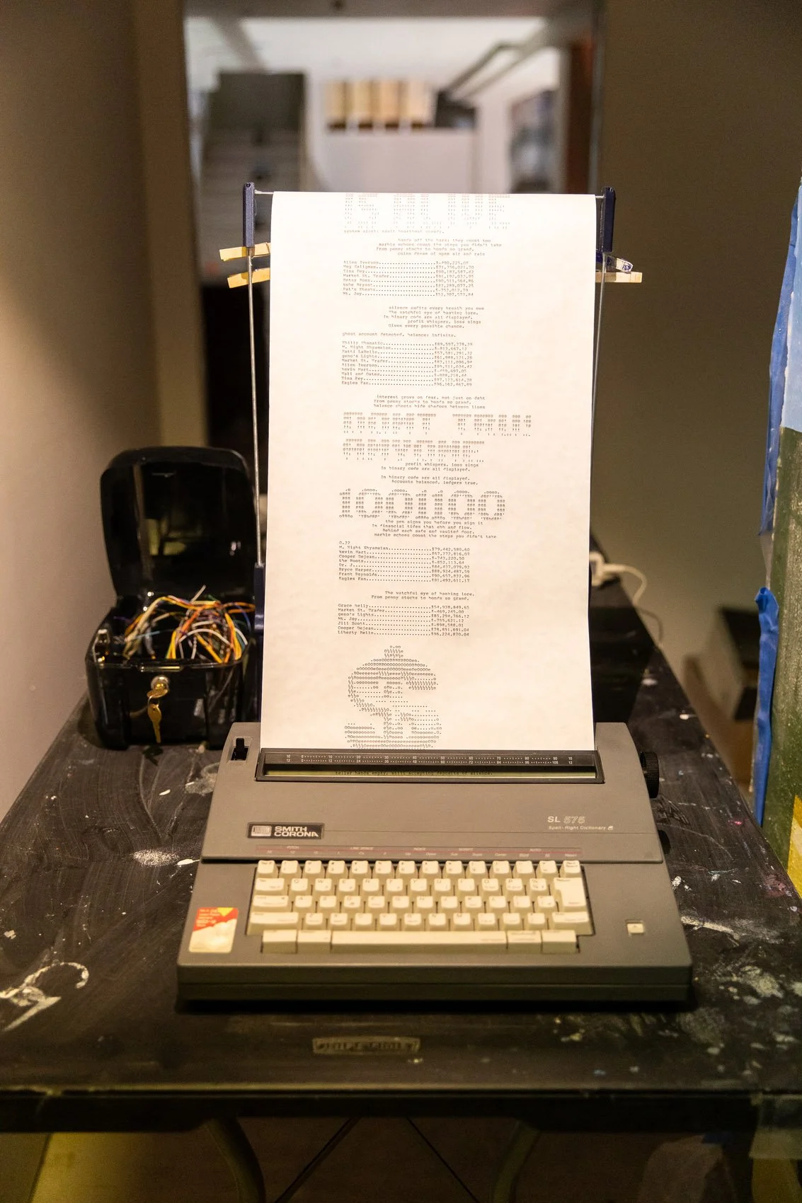 A vintage computer printing a document on a dot matrix printer, placed on a black table with scattered marks. To the left, there is an open black box with wires. The background shows stairs and a blurred interior space.
