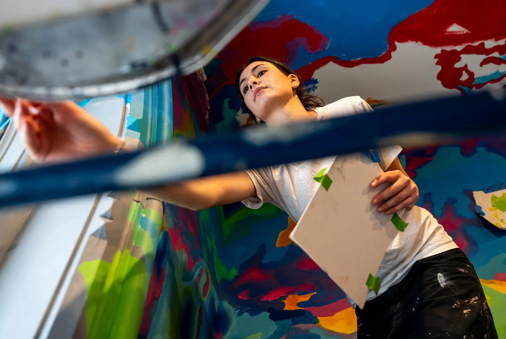 Woman painting a colorful mural on a wall, viewed from below through a railing.