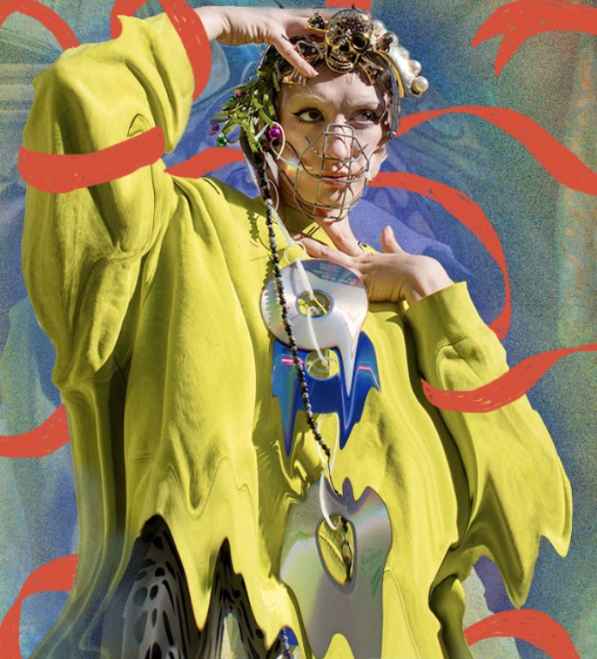 A person wearing a bright yellow oversized hoodie, with elaborate jewelry and accessories, including a face mask made of metal wires, beads, and decorative elements, posing against a colorful background with red accents.