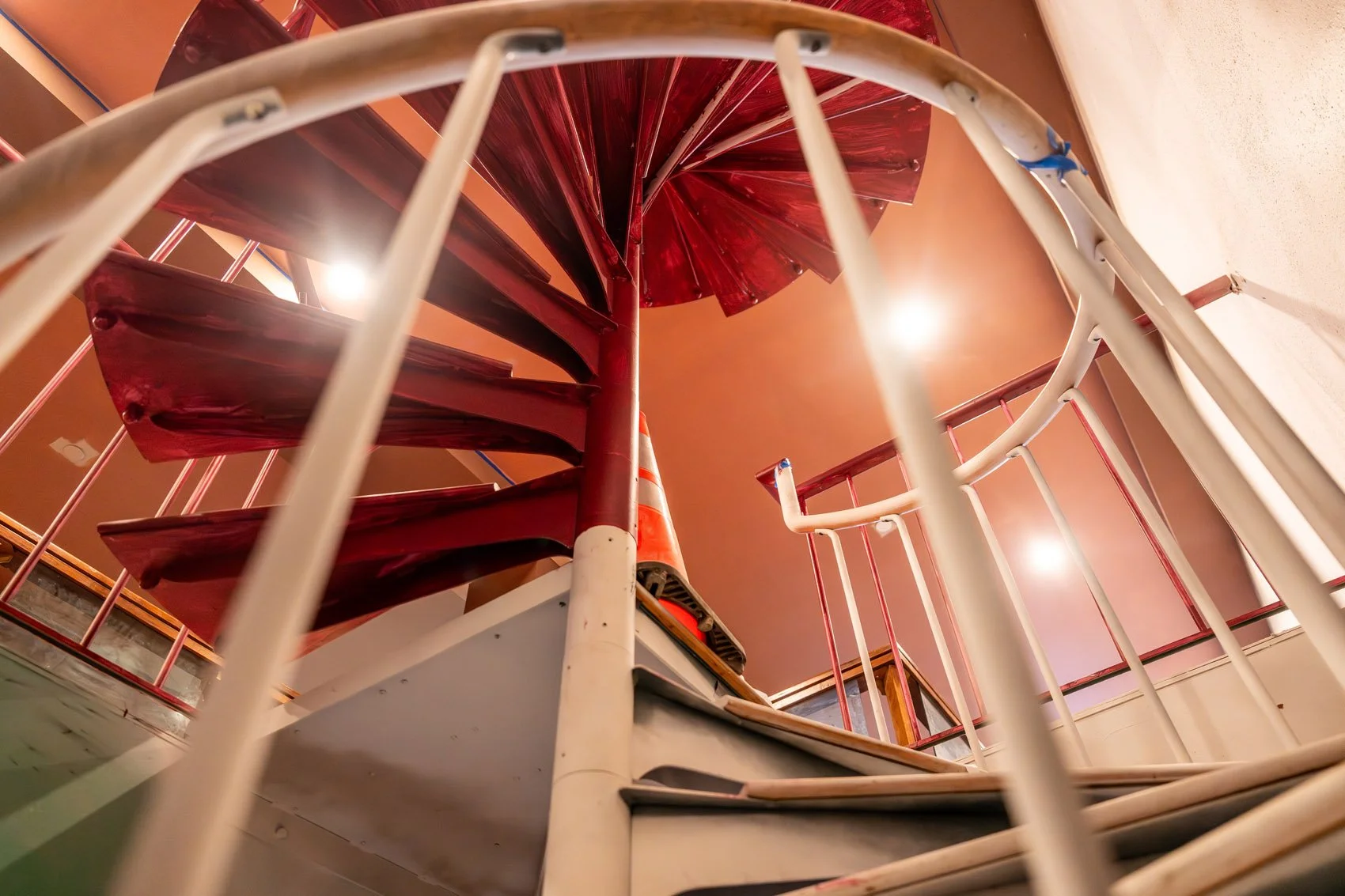 View from the bottom of a red spiral staircase with white railings, looking upward toward the ceiling and the spiral structure.
