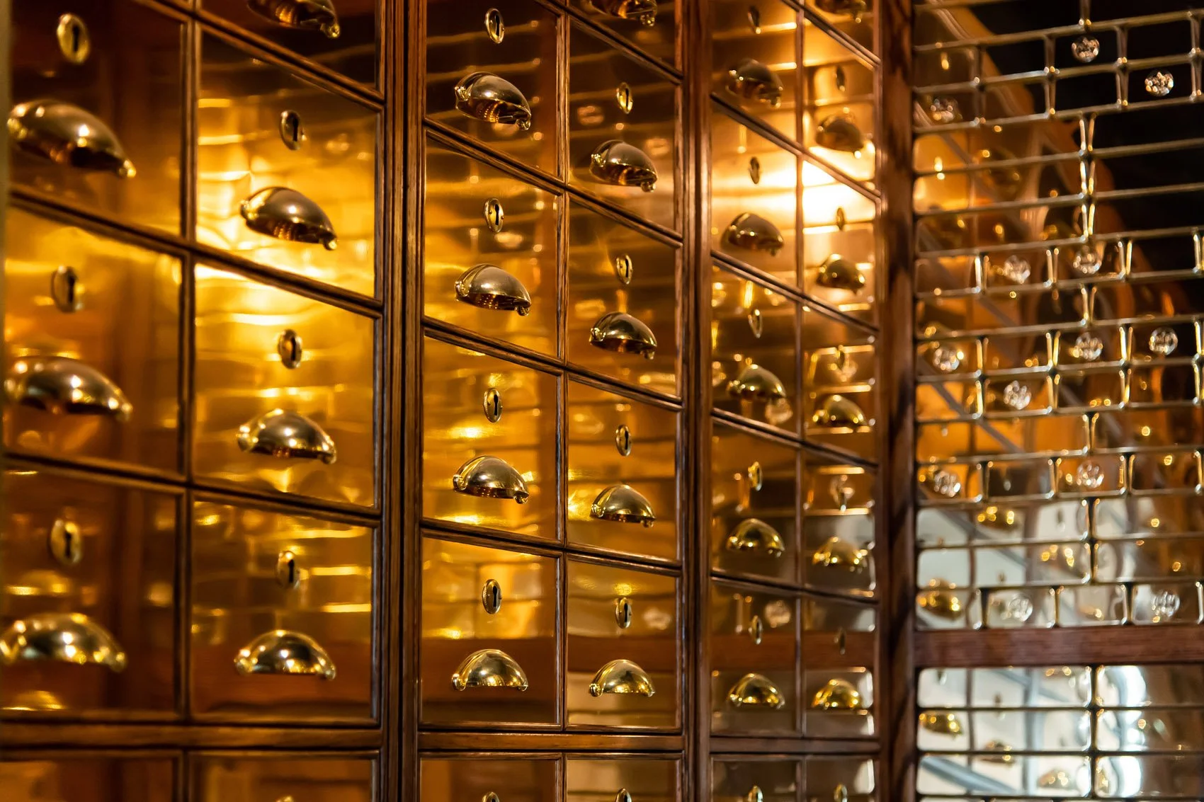 A wall of small, shiny, brass-colored drawers with metallic handles, set in a wooden frame, with reflections of warm lighting.