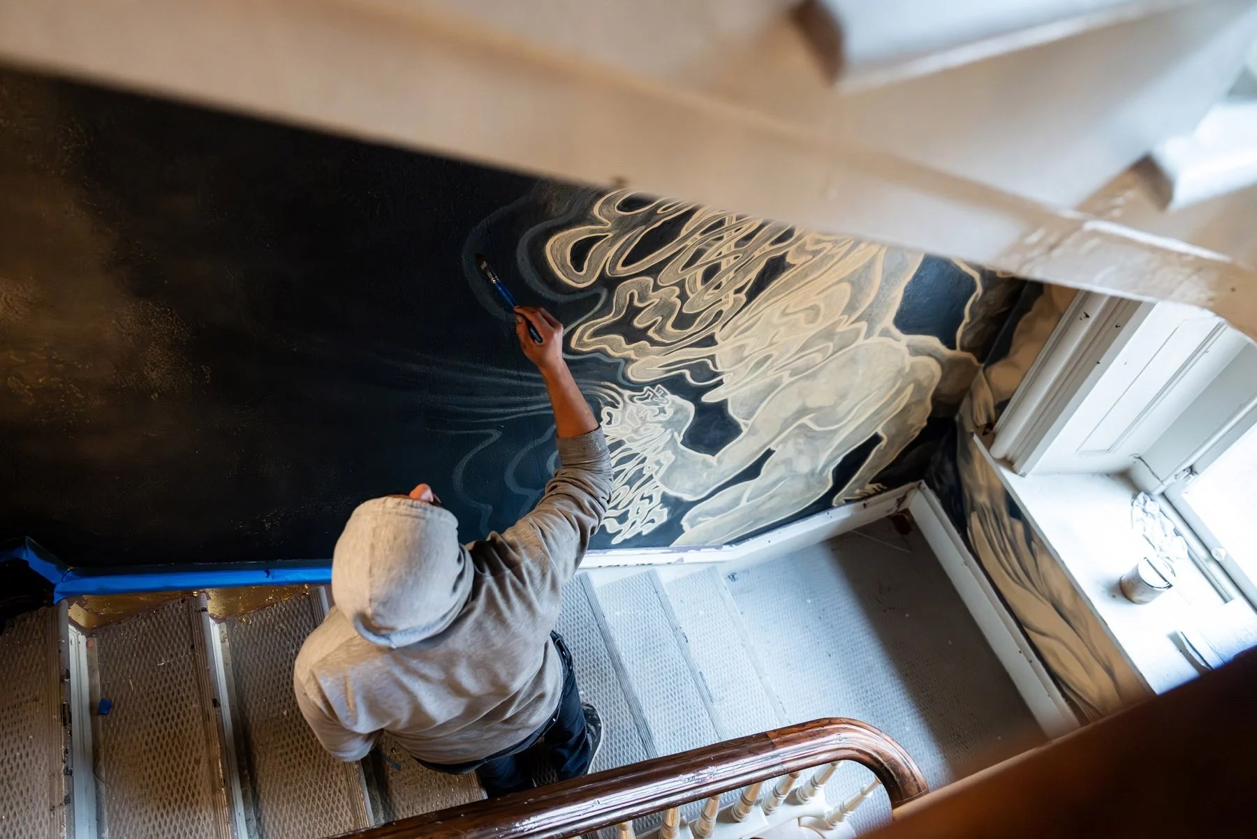 Person painting a mural on a staircase wall using a paintbrush, with swirling white and black patterns.