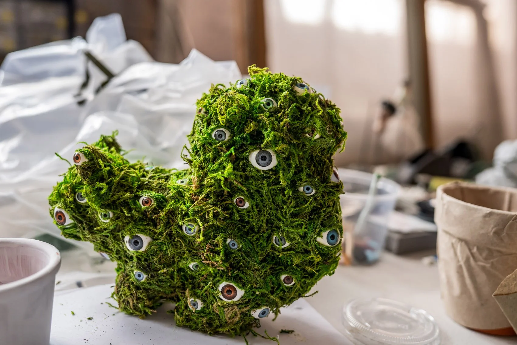 A moss sculpture shaped like a head with multiple plastic eyes attached, placed on a work surface in a workshop.