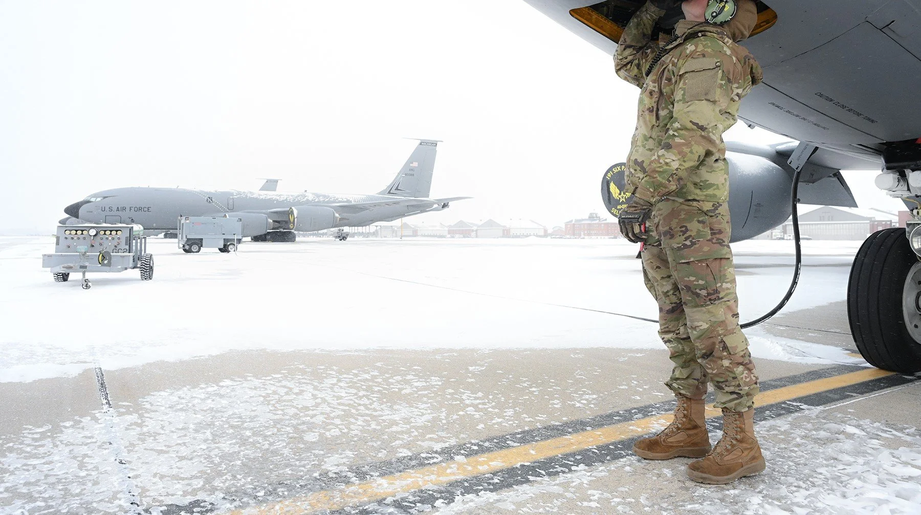 A military crew member in camouflage uniform and boots standing under the wing of a military aircraft, refueling it on a snowy tarmac with another similar aircraft in the background.