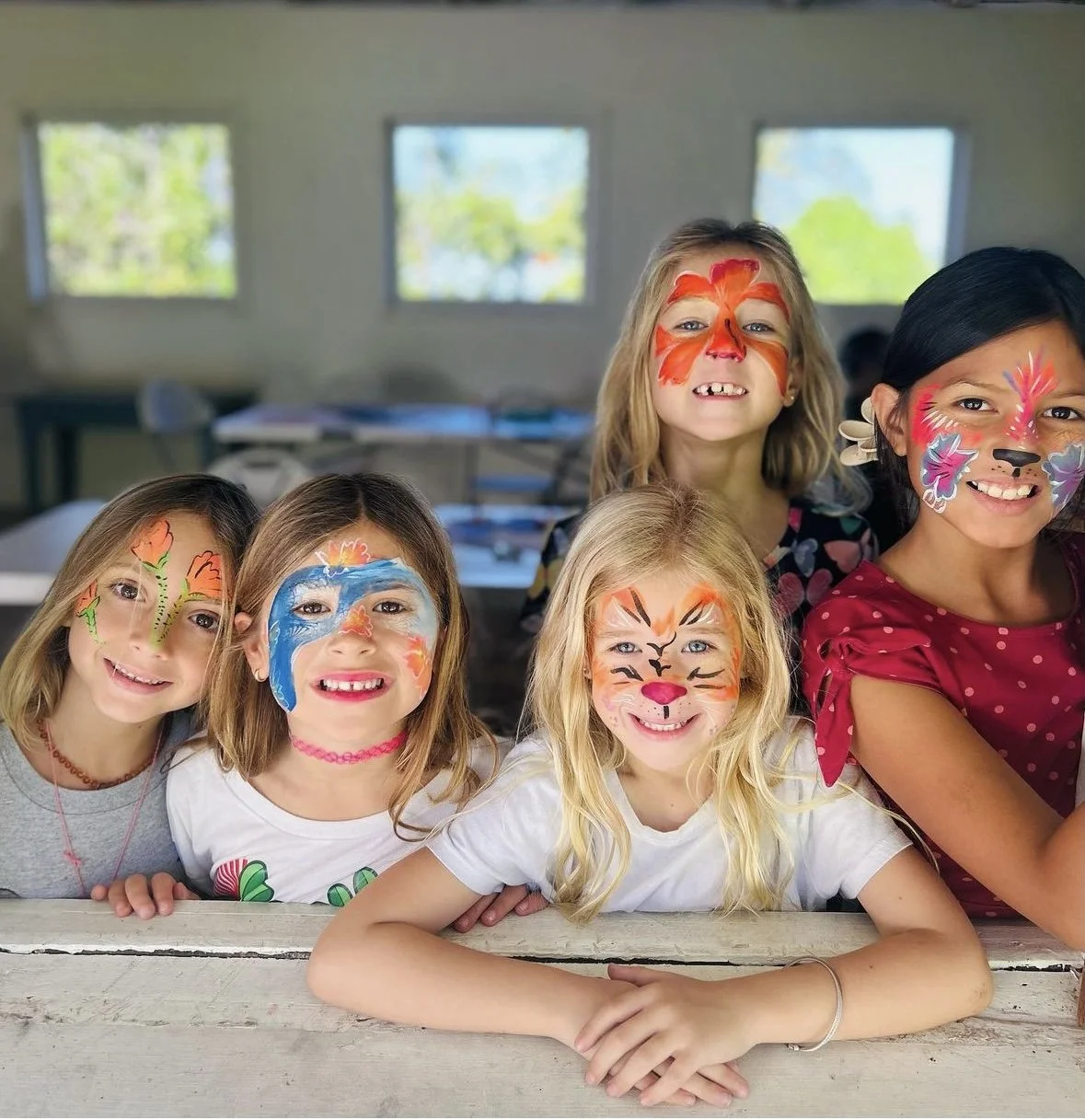 Six young girls with face paint, smiling, indoors in a classroom setting.