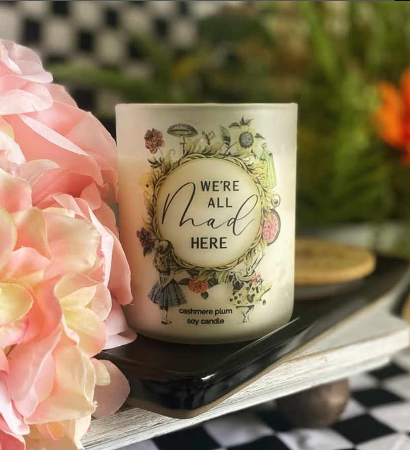 A white candle with floral and girl illustrations, labeled 'We're all mad here,' placed on a black tray next to pink flower petals.