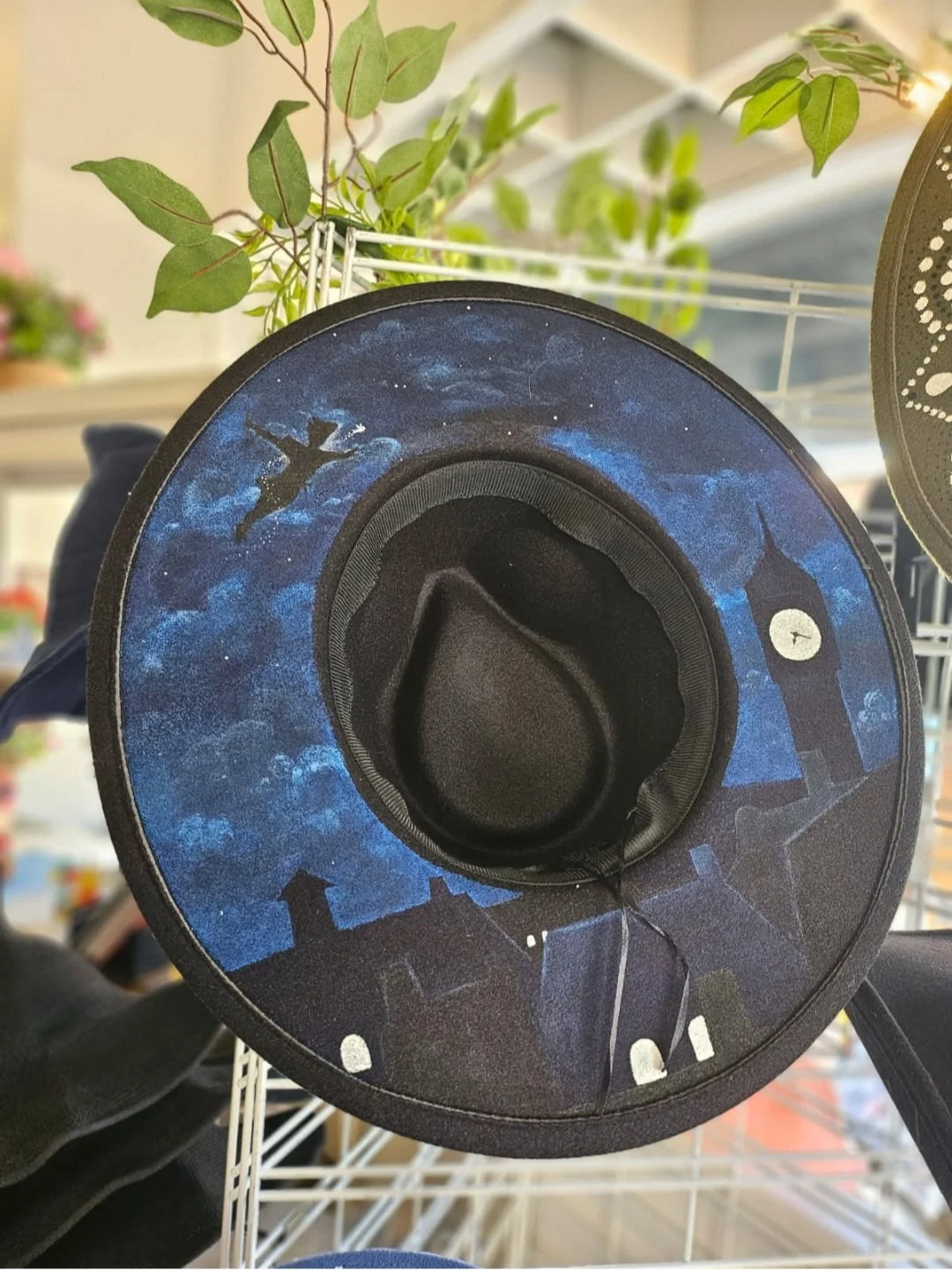 A hat with a night sky design, featuring a silhouette of Peter Pan flying, a clock tower, and buildings, displayed on a wire shelf with green leaves in the background.