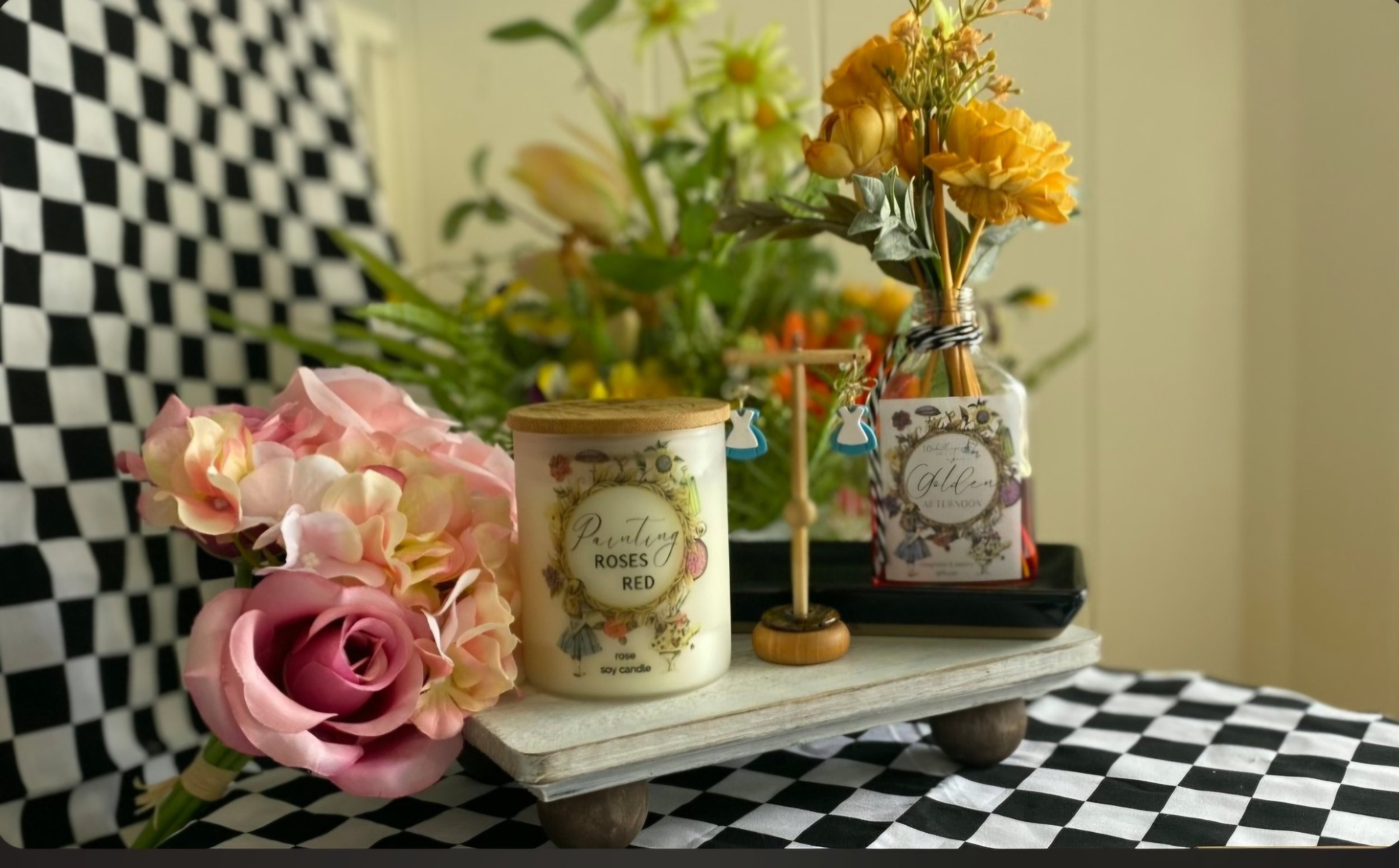 A decorative arrangement on a wooden tray featuring pink and peach artificial roses, a white candle with floral design labeled 'Painting Roses Red,' a small flag, and a glass jar filled with yellow flowers, all set against a background of green foliage and a checkered black-and-white fabric.