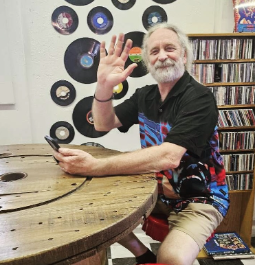 An older man with gray hair and a beard waving while sitting at a wooden table in front of a white wall decorated with vinyl records. He is smiling and holding a smartphone.