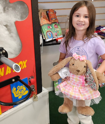 A young girl with long brown hair smiling and holding a teddy bear dressed in a pink checkered dress, standing near an arcade game machine.
