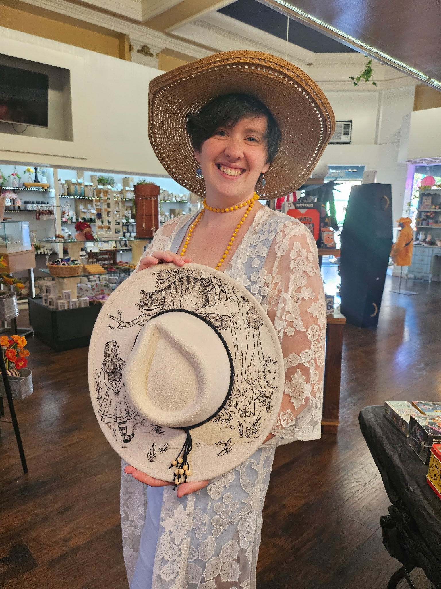 A woman smiling inside a store, wearing a large woven straw hat, a yellow beaded necklace, and a white lace dress. She holds a decorative hat with a drawing of a girl and a cat on it.