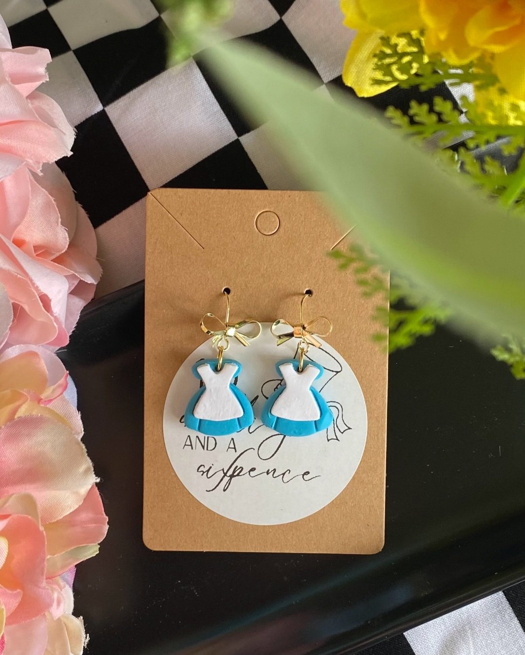 Earrings shaped like dresses with blue skirts and white bodices, gold bows at the top, displayed on a craft card with text, surrounded by pink and yellow flowers and greenery.
