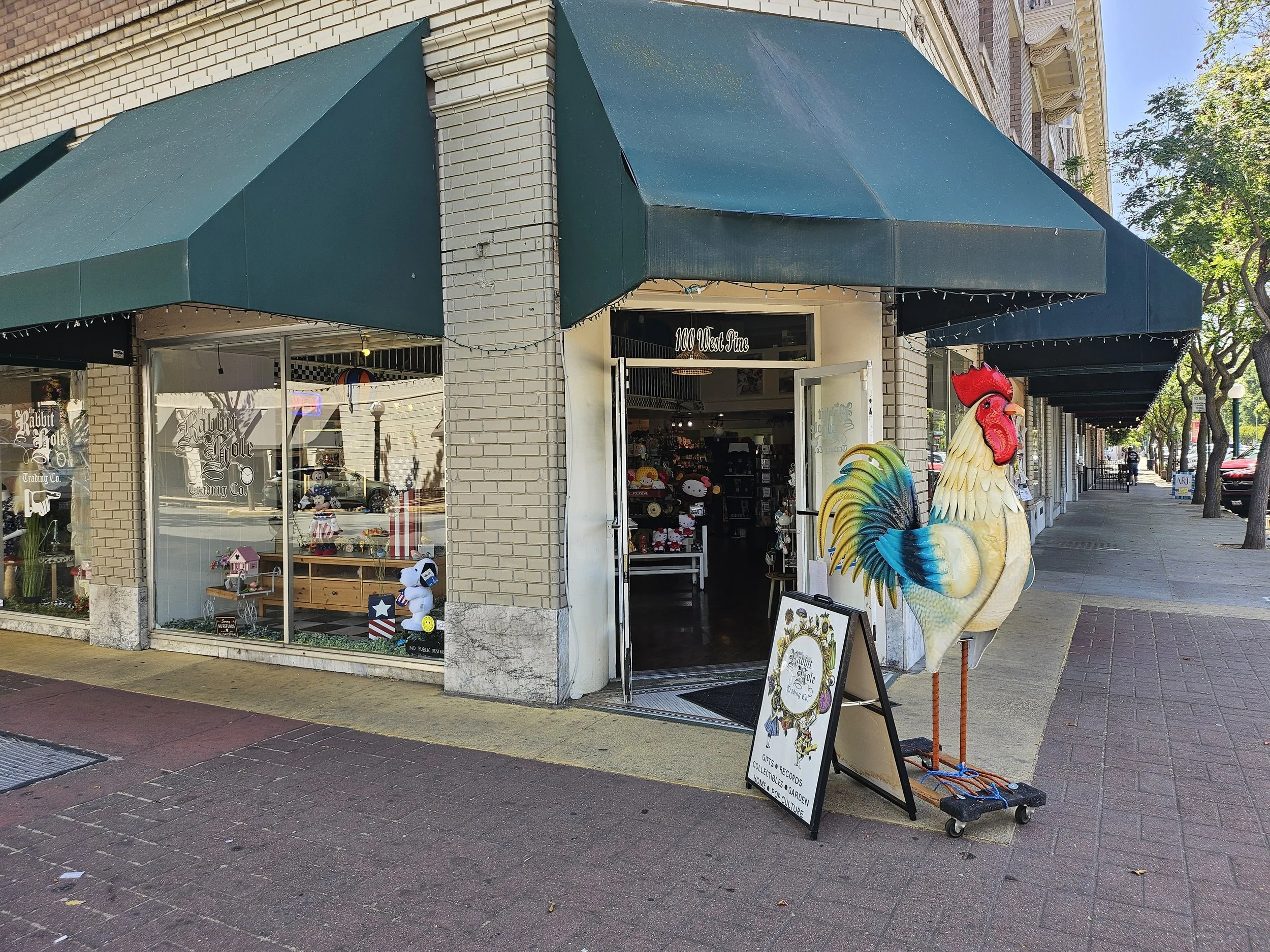The exterior of a storefront with green awnings on a city sidewalk, decorated with a large rooster figure and a signboard displaying gift and record store information.