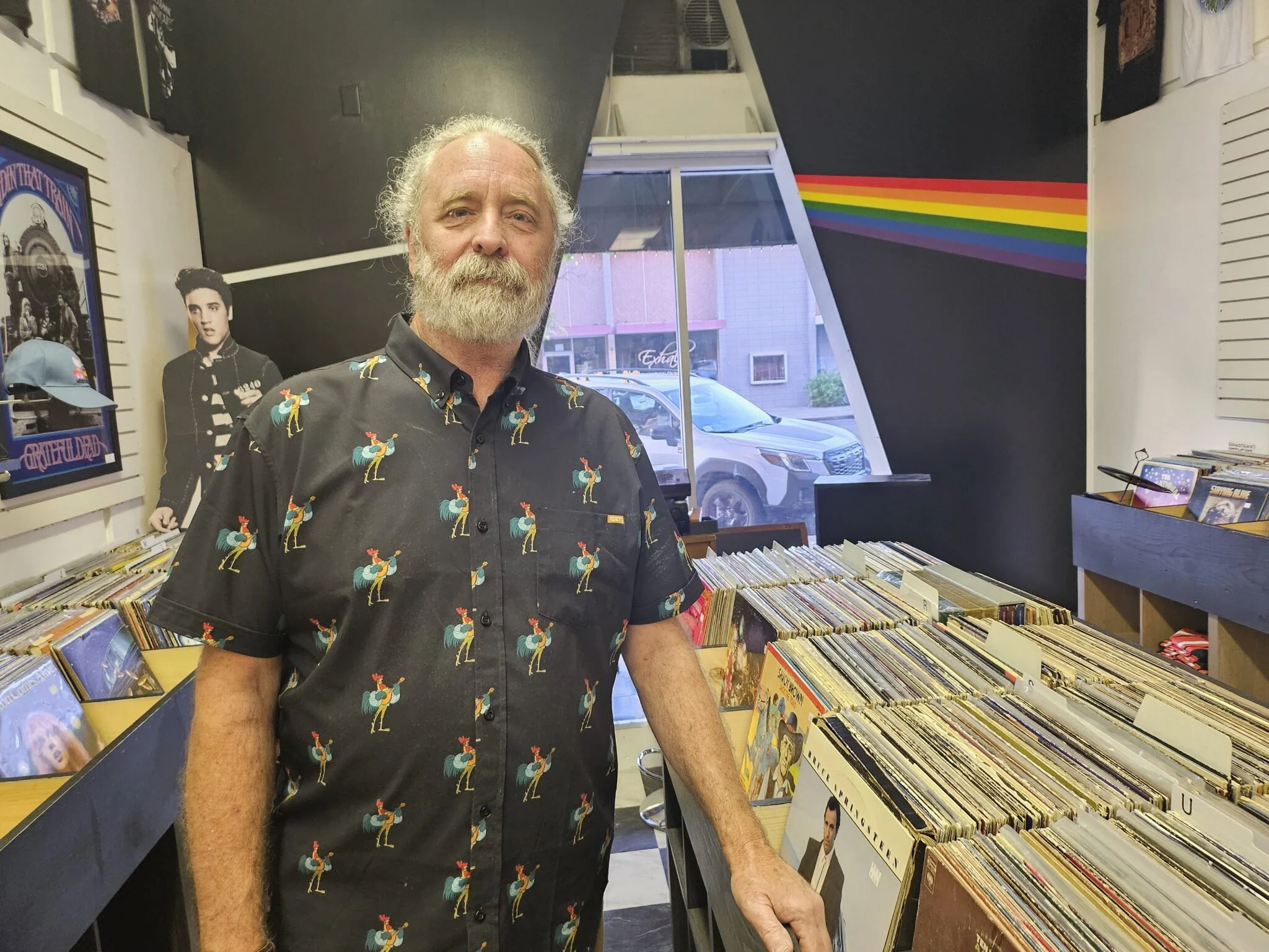 A man with a gray beard and long gray hair standing inside a record store, wearing a black shirt with a colorful rooster pattern. The store has records displayed in bins and posters on the wall, including a black-and-white picture of Elvis Presley.